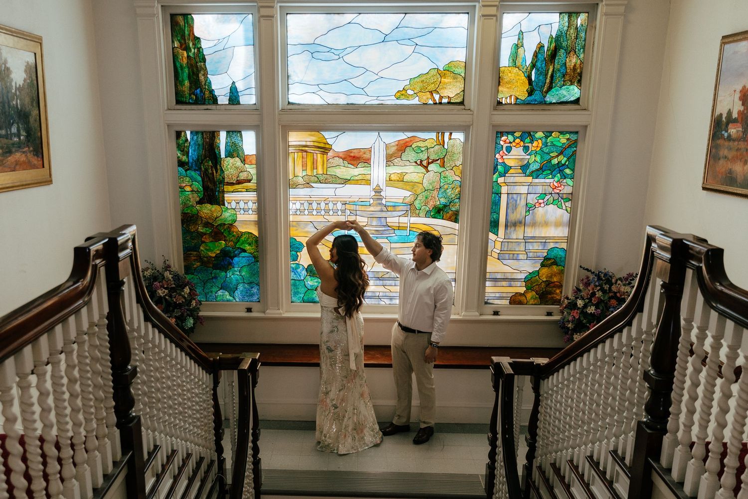 Bride and groom dancing together on staircase landing before vibrant stained glass window in historic venue.