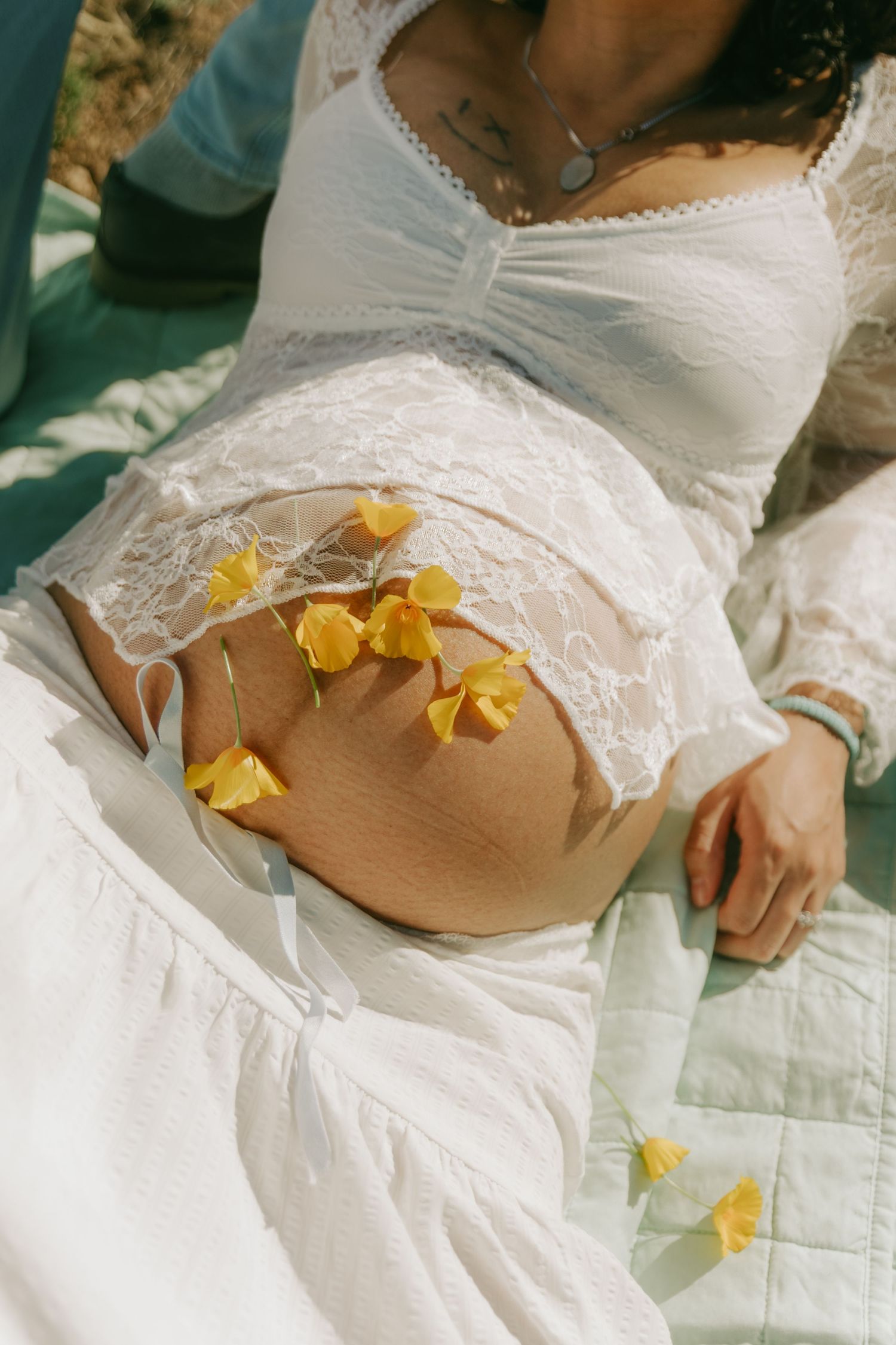 Close-up of pregnant belly adorned with small yellow wildflowers, woman wearing white lace dress outdoors.