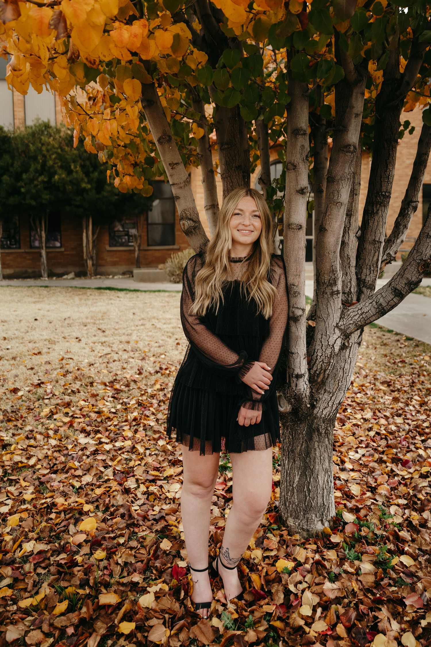 Smiling blonde woman in black dress standing under colorful autumn trees on campus.