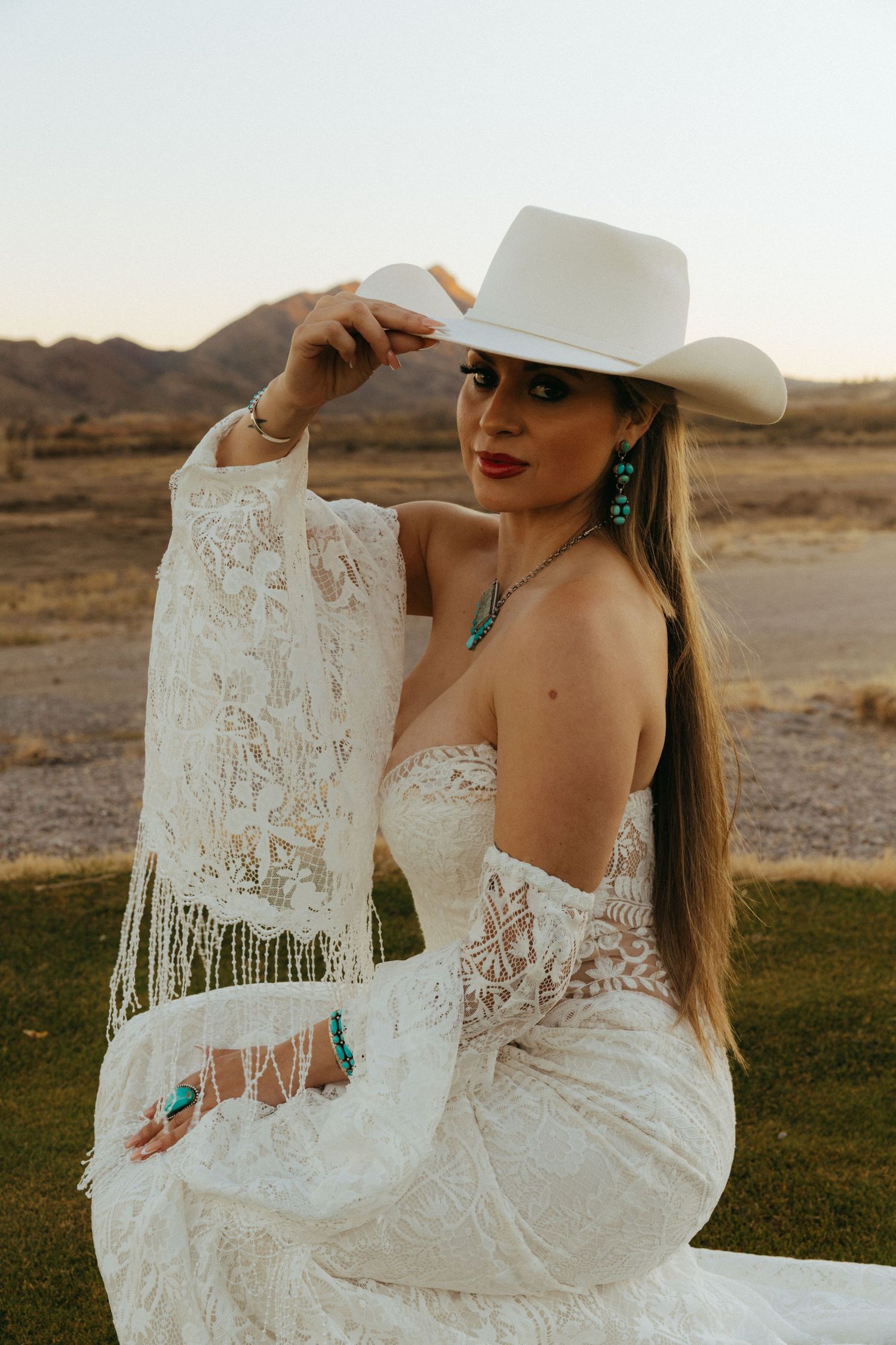 Woman in white lace wedding dress and cowboy hat poses in desert landscape at sunset.