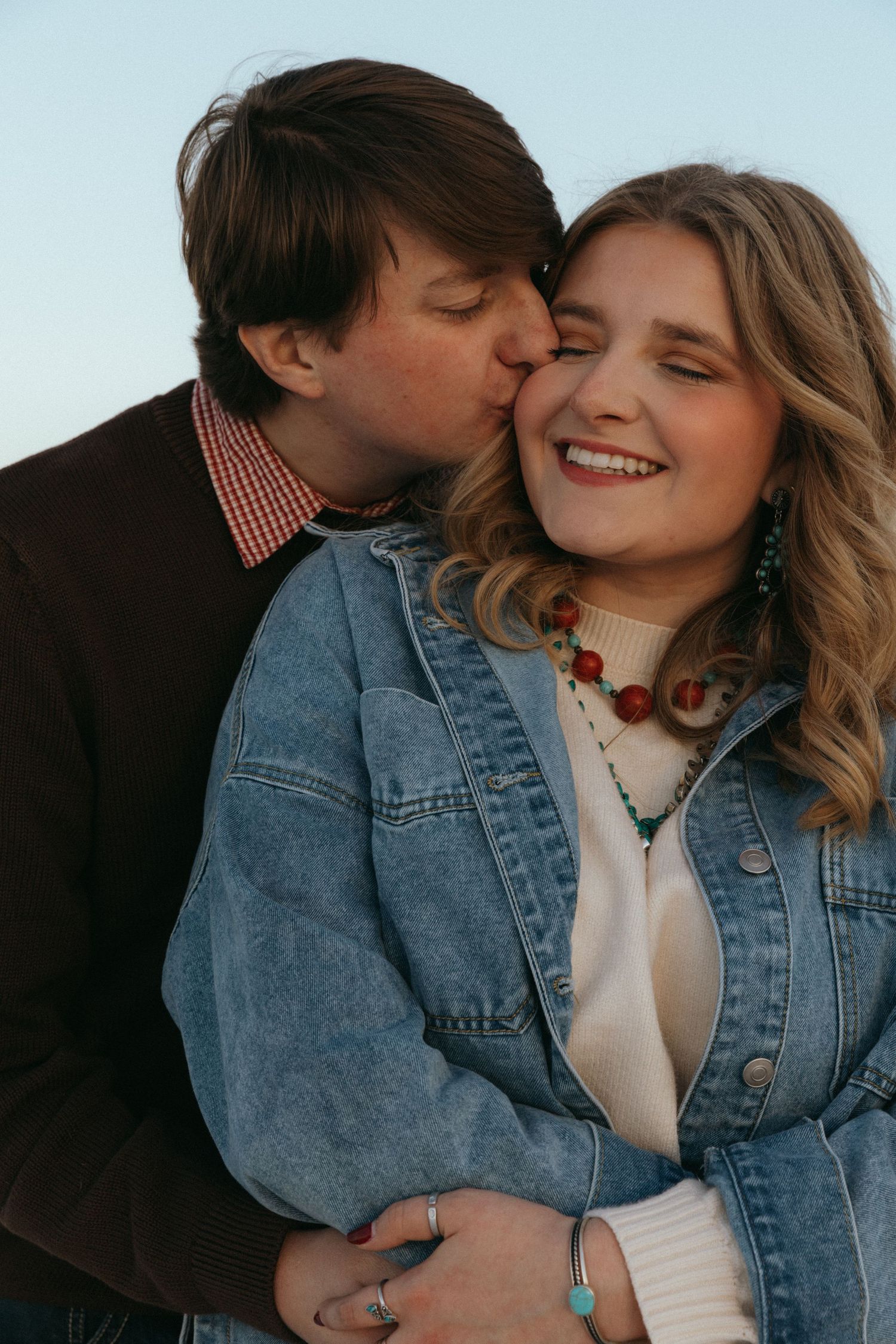 A man kisses a smiling woman wearing a denim jacket and colorful beaded necklace outdoors.