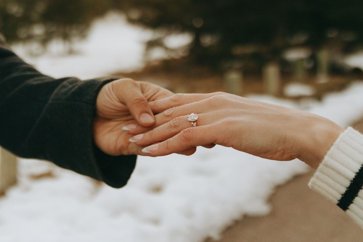 Two hands touching with a diamond engagement ring on a womans finger in a snowy winter outdoor setting.