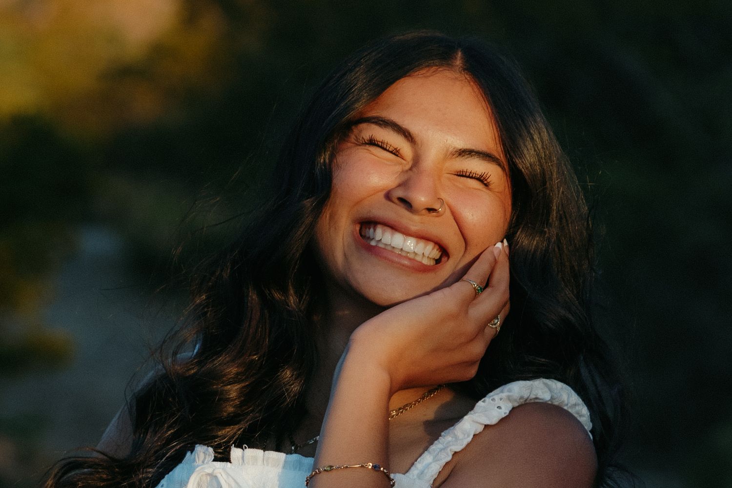 Young woman with long dark hair laughing joyfully, hand on cheek, wearing a white top and bracelet outdoors.