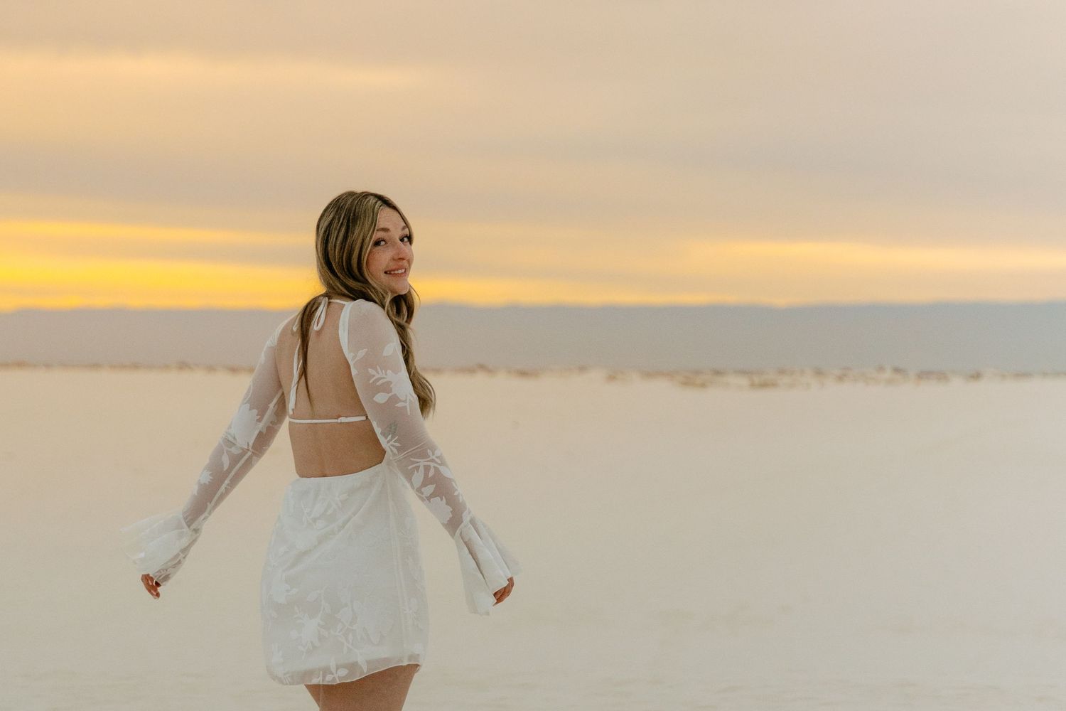 Smiling woman in white backless lace dress turns toward camera on white sand at sunset.