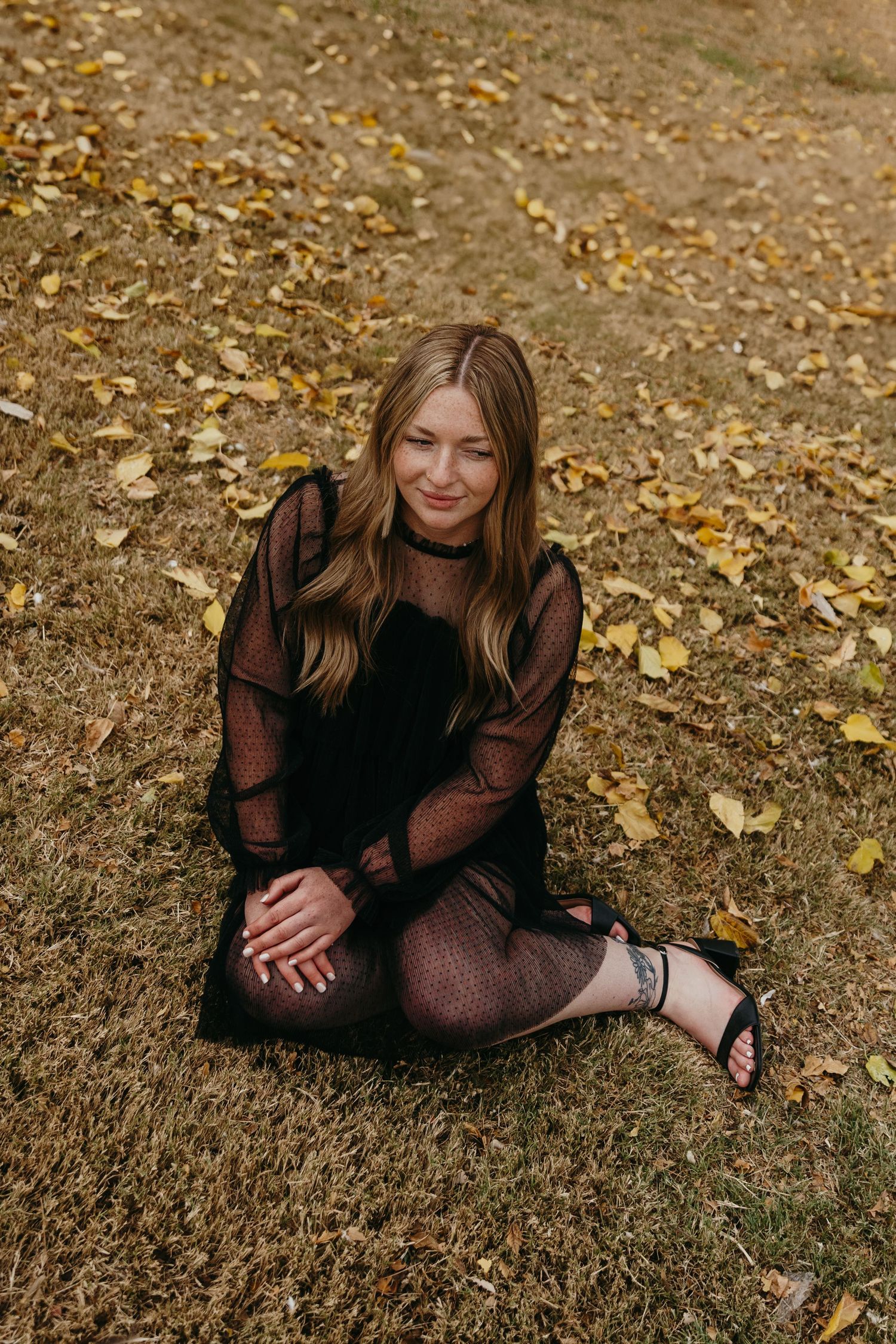 Young woman in black sheer dress sitting on grass surrounded by fallen autumn leaves.