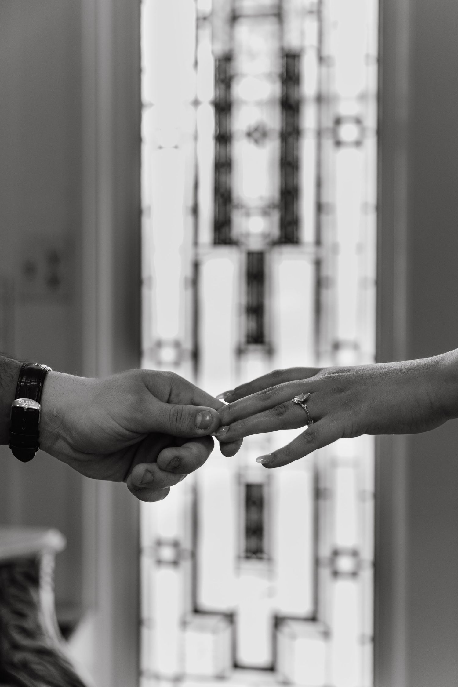 Black and white photo of a couple exchanging wedding rings in front of a stained glass window.