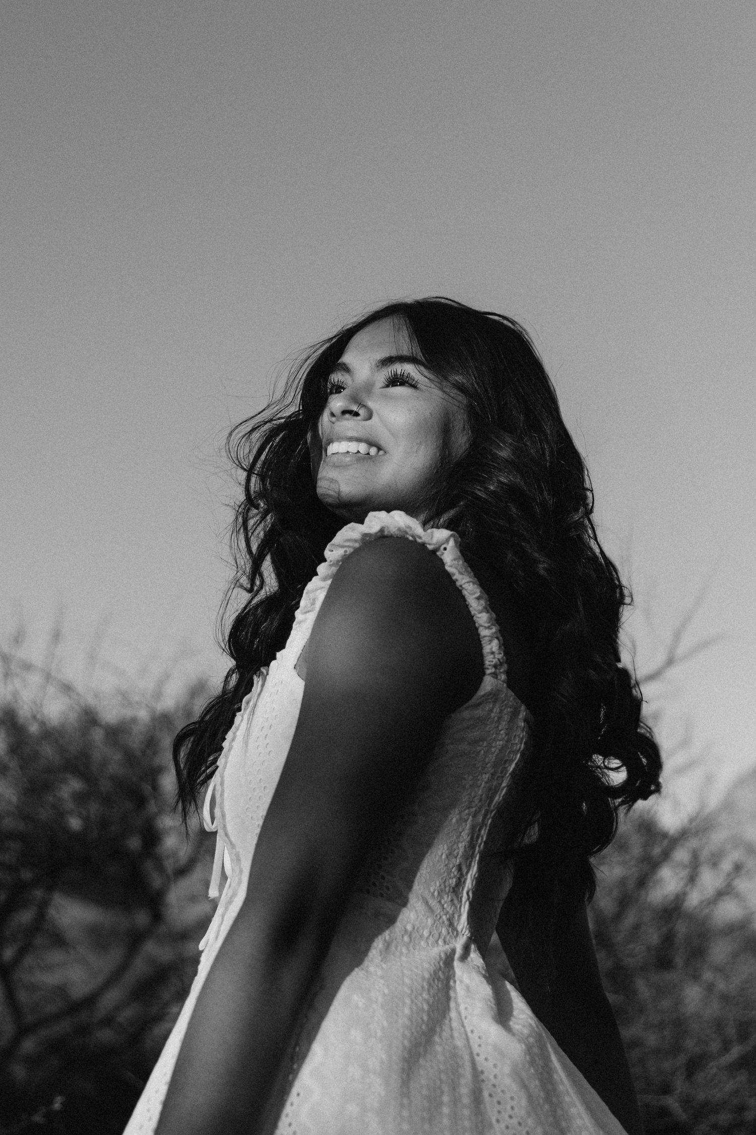 Black and white portrait of smiling young woman with long wavy hair in white dress against open sky.