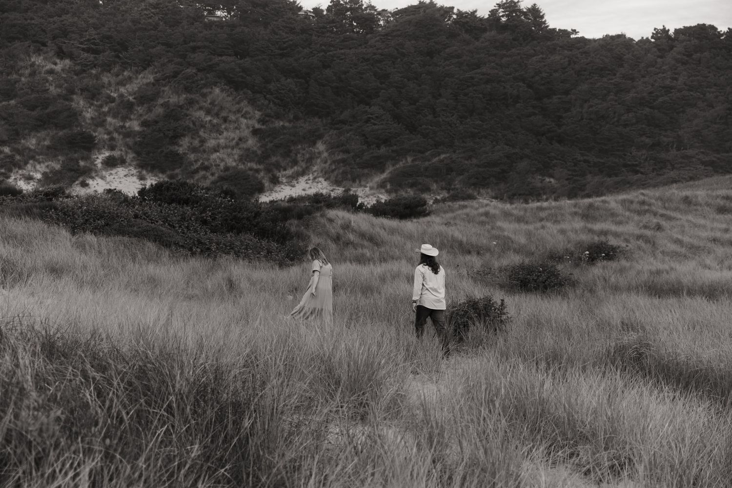 Two people walking through tall grass in a black and white landscape with rolling hills and dense trees.