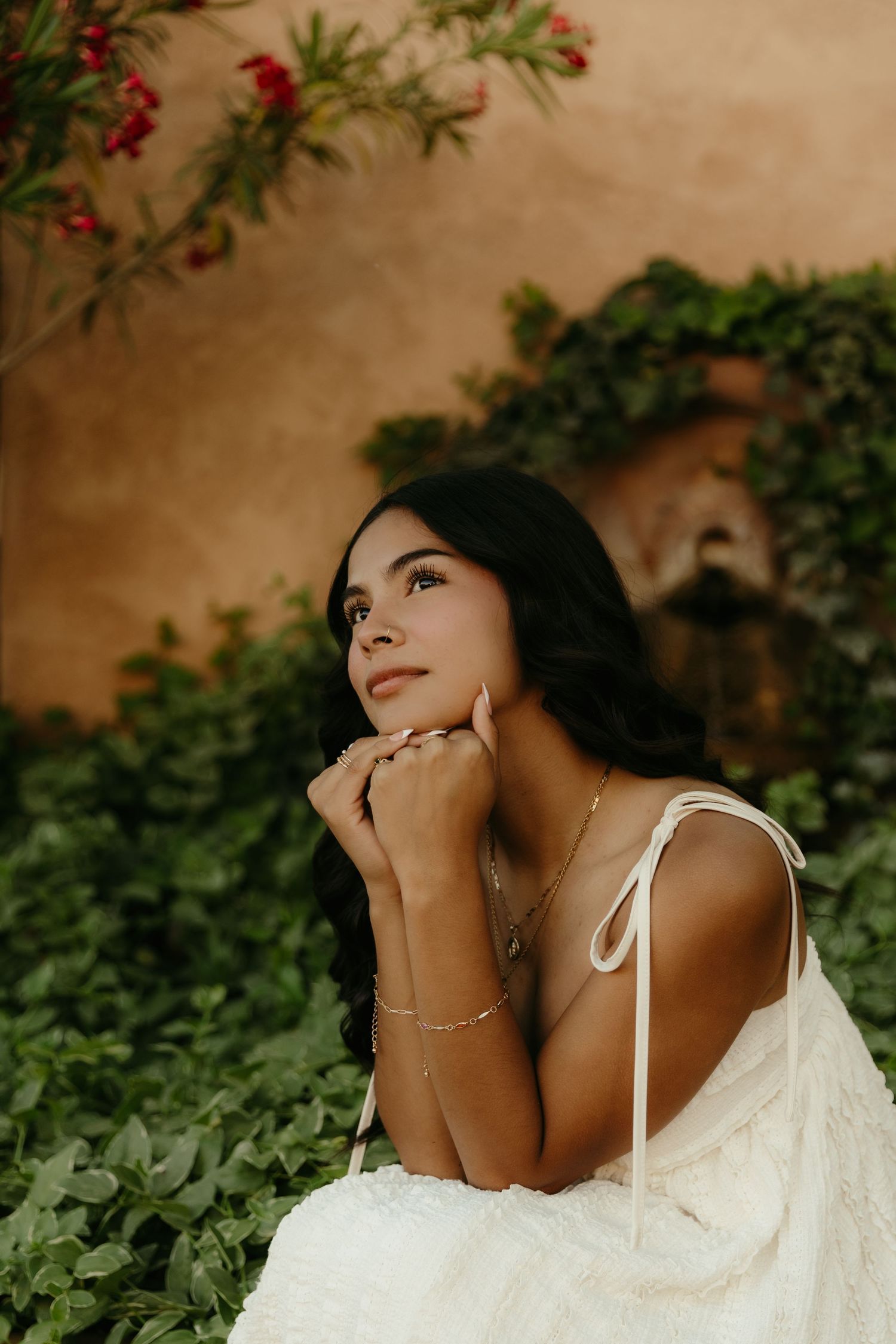 A woman in a white dress sits dreamily among lush green ivy, resting her chin on her hands near a terracotta wall.