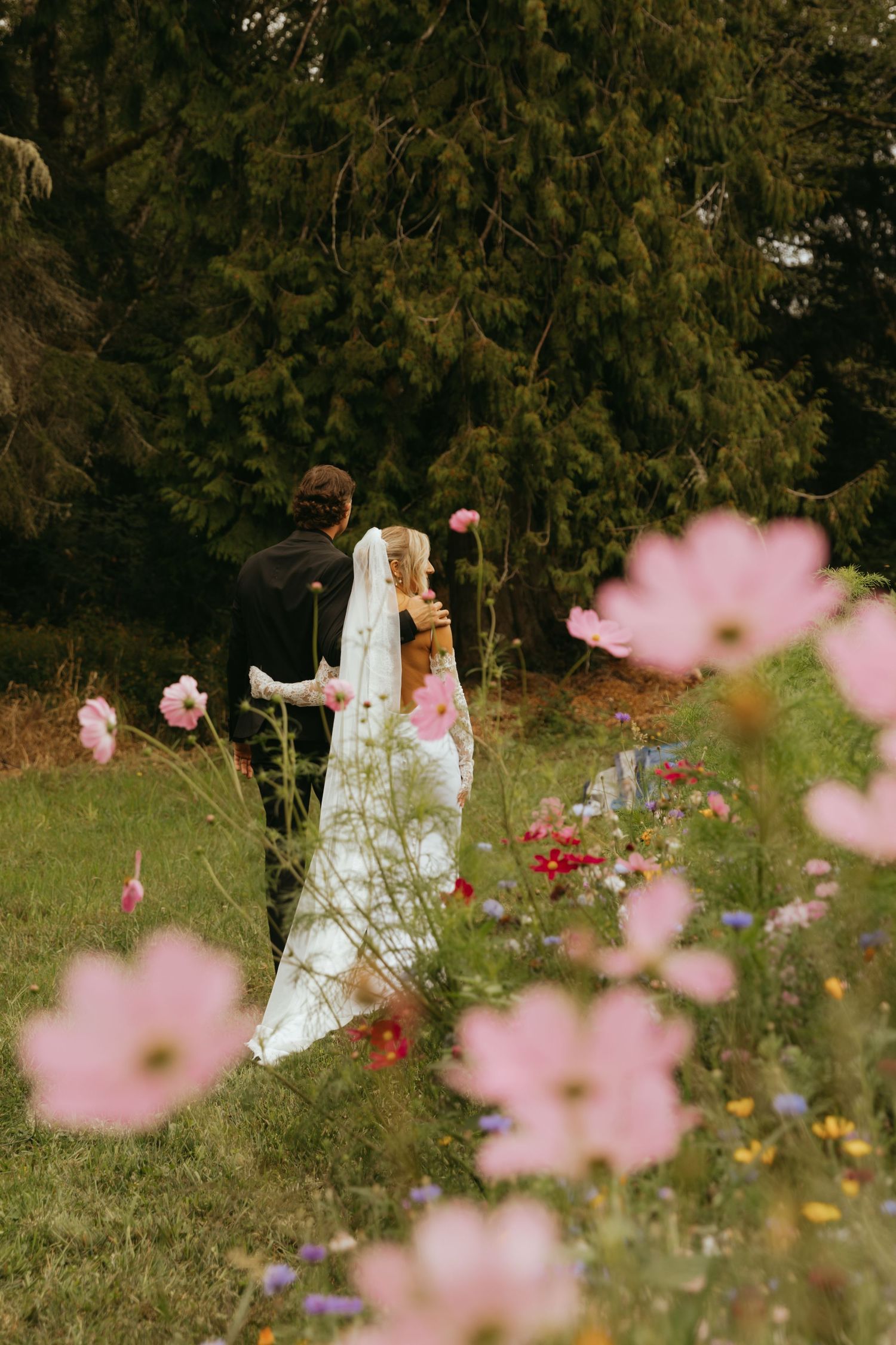 Bride and groom walking through a wildflower meadow with pink cosmos blooms in foreground and evergreen trees behind.