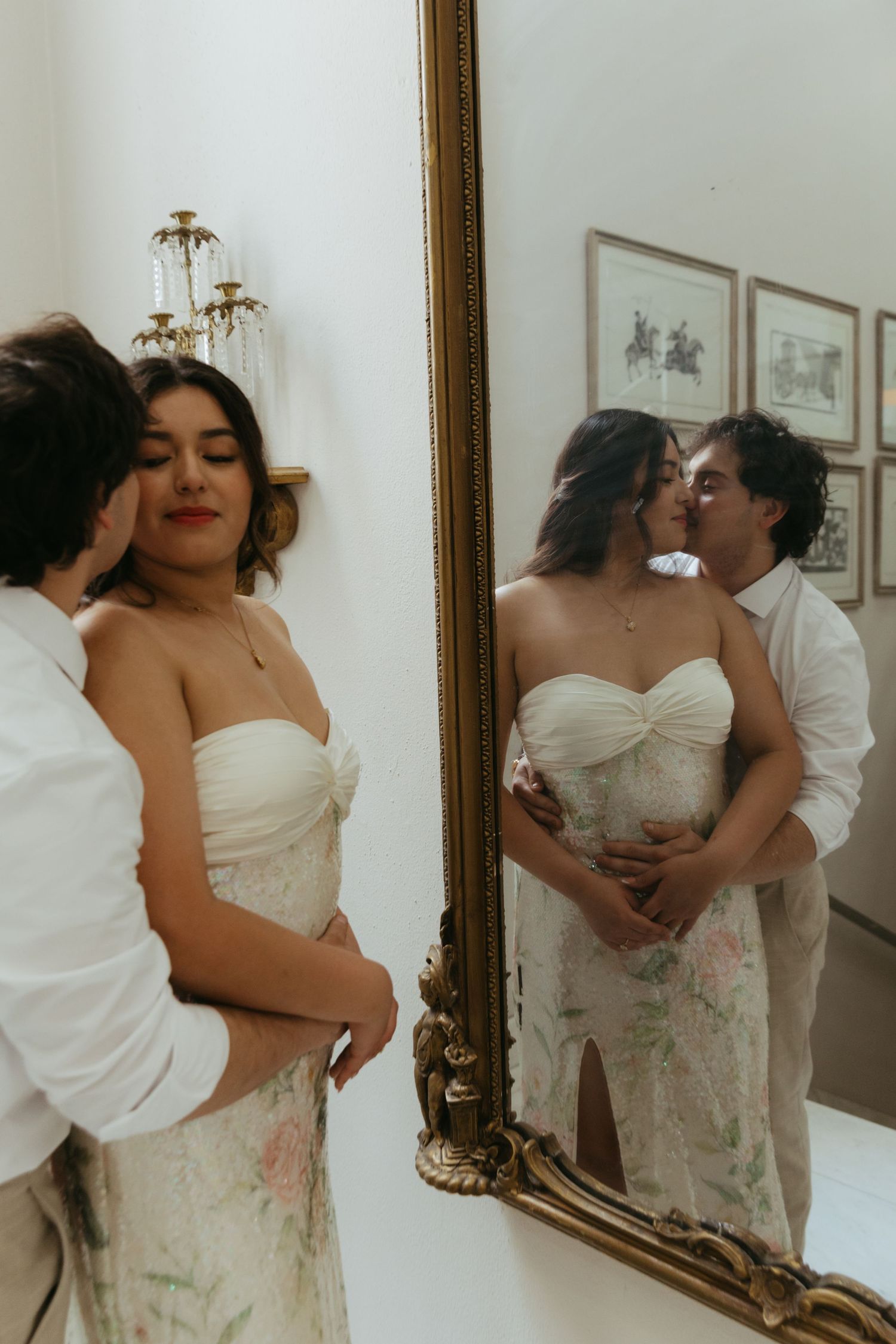Couple in wedding attire embracing before a gold-framed mirror, reflection showing a romantic kiss.