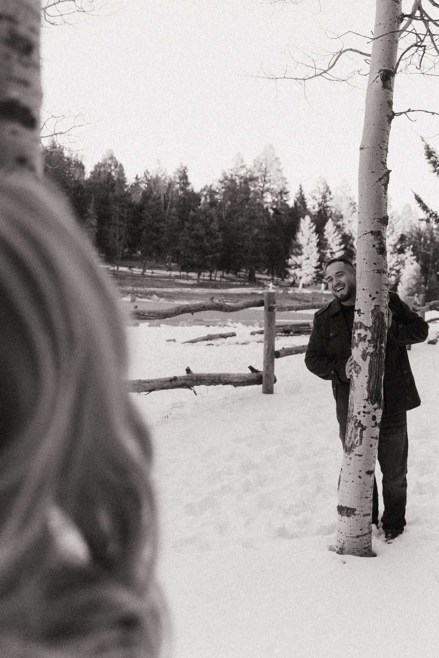 Black and white winter photo of a smiling man leaning against a birch tree in a snowy landscape with evergreens.