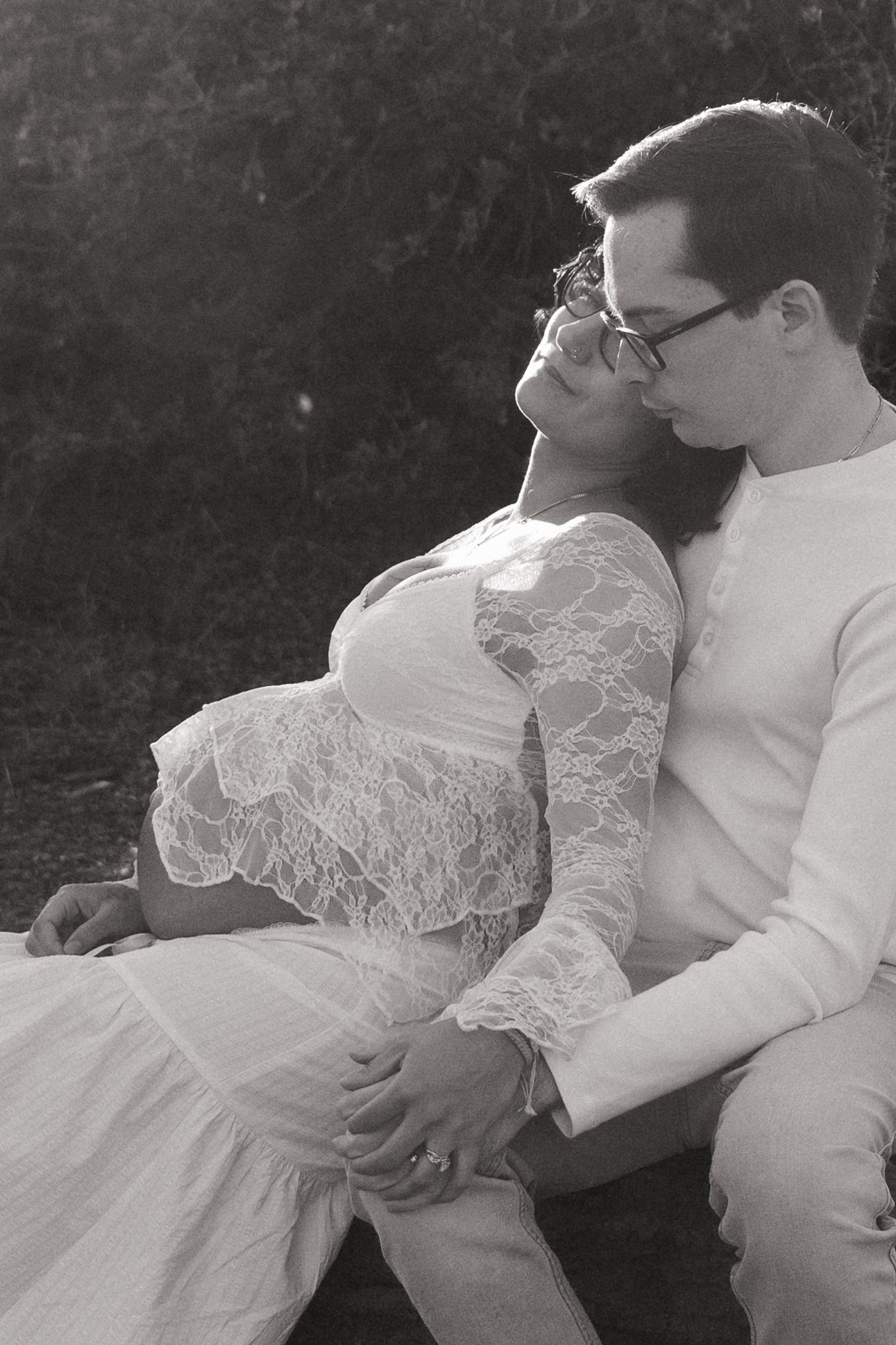 Black and white maternity photo of a couple sitting outdoors, woman in lace dress, man in white outfit kissing her cheek.