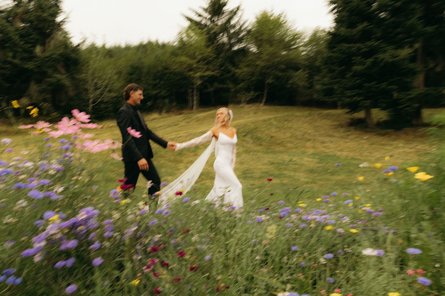 A bride in white gown and groom in black suit run hand-in-hand through a vibrant wildflower meadow.