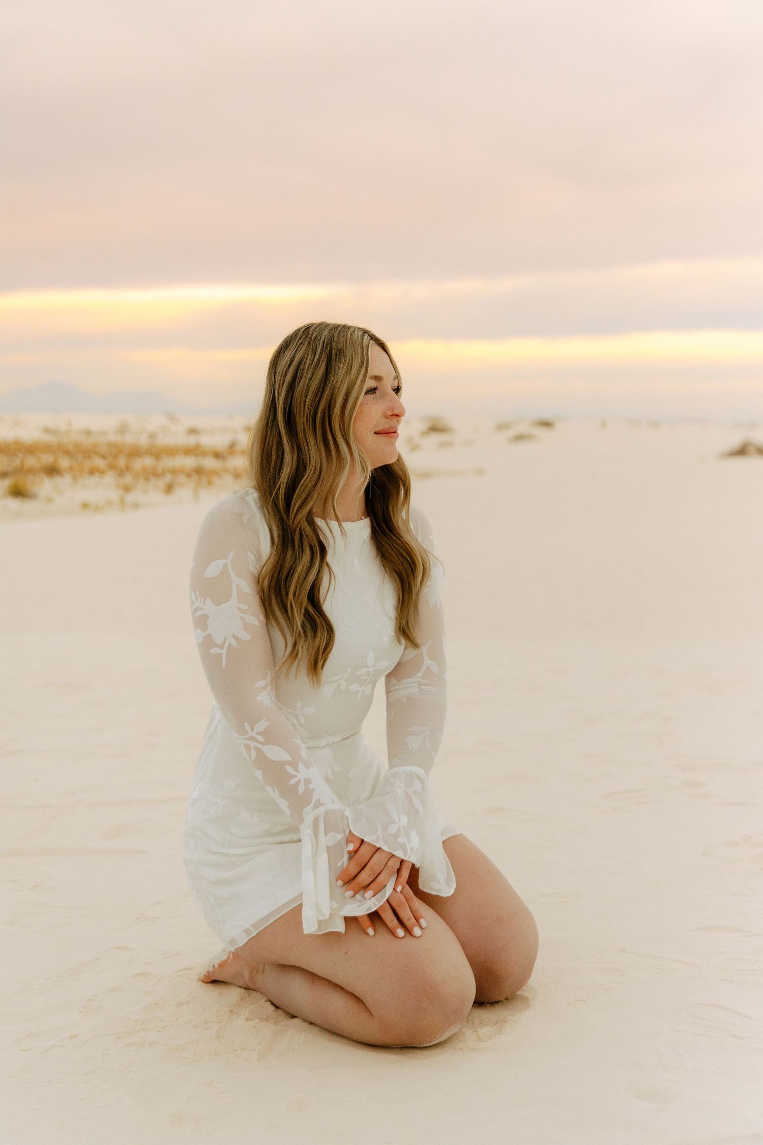 Blonde woman in white lace long-sleeve dress kneels on white sand gazing at colorful sunset sky.