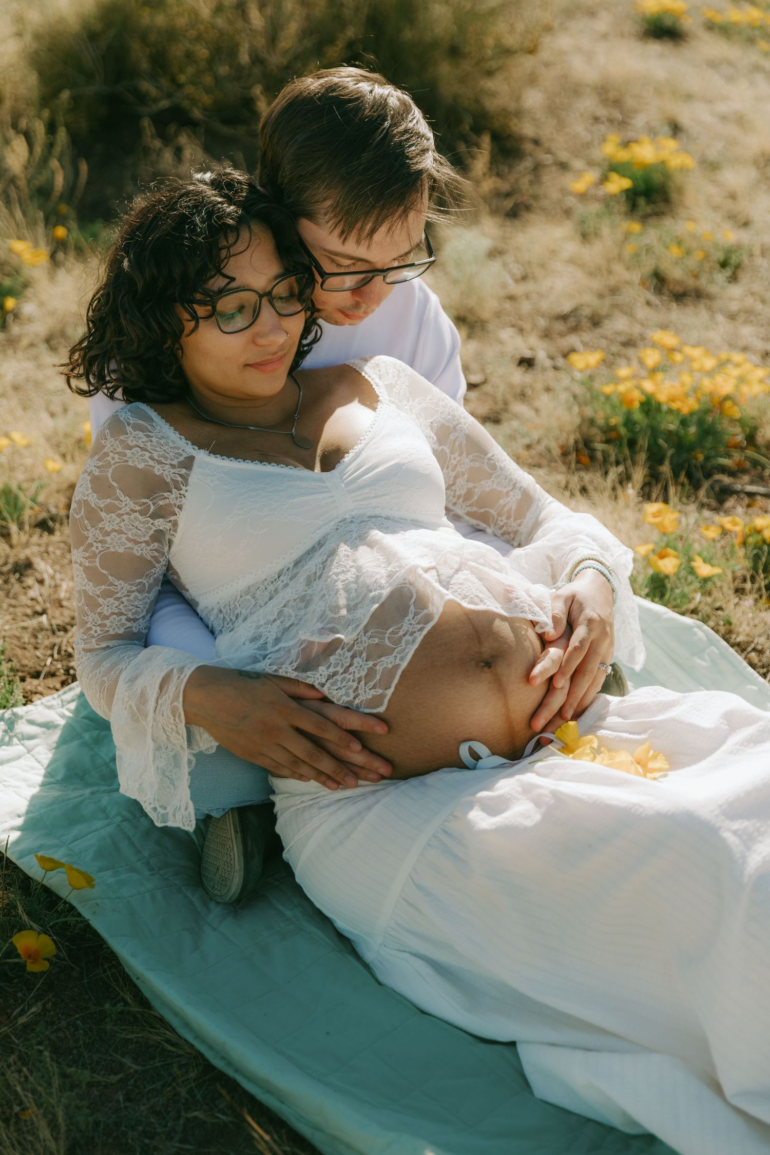 Couple sharing tender moment outdoors, partner embracing pregnant woman in white lace dress while sitting in flower field.