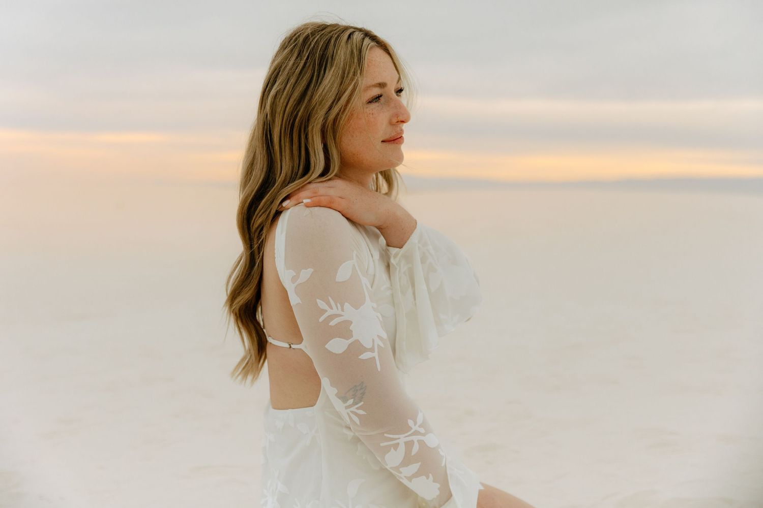 Blonde woman in elegant white floral lace long-sleeve dress gazes away on white sand at sunset.
