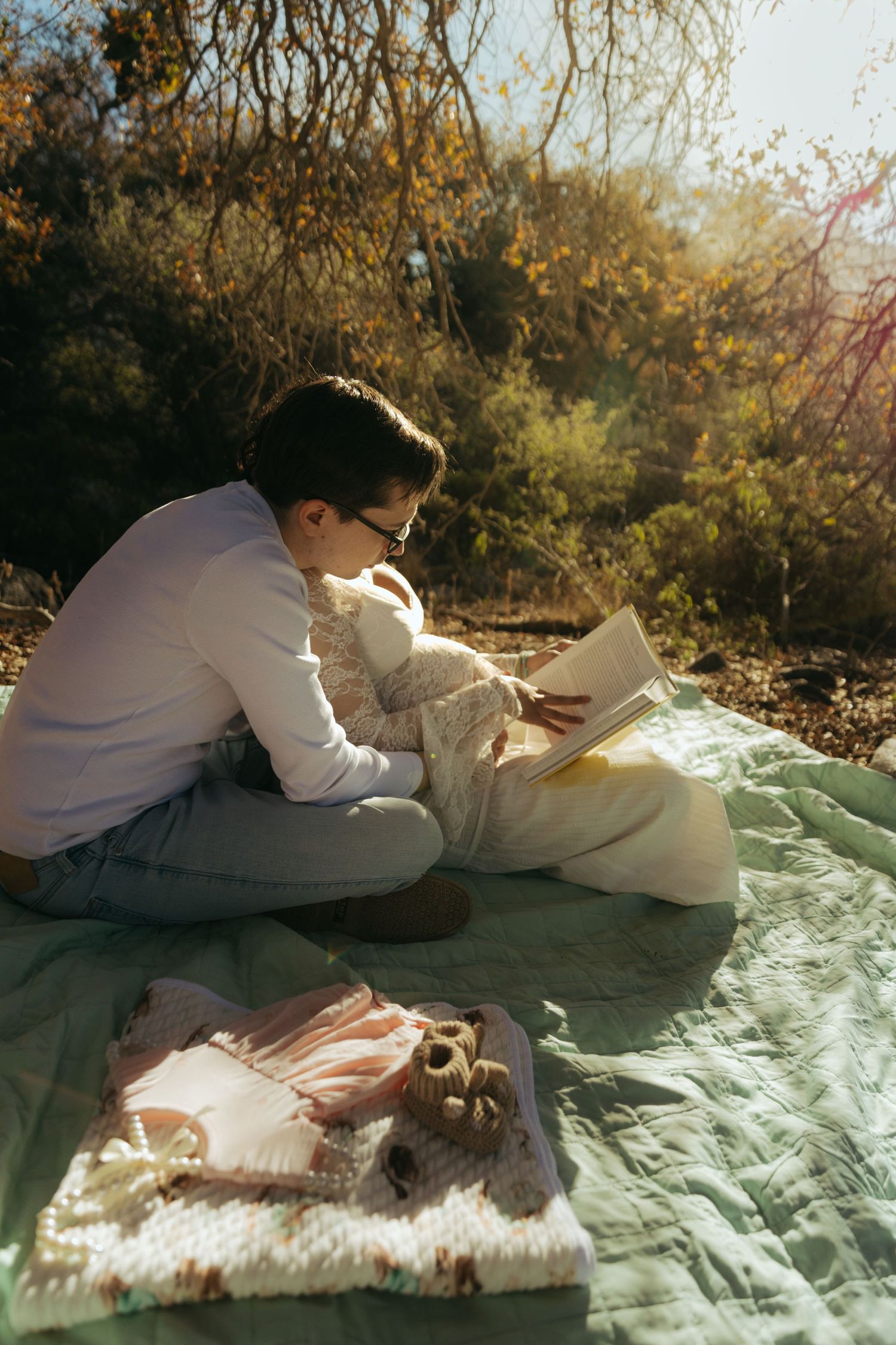 Father-to-be reads baby book on mint blanket with infant clothes and booties in sunlit natural outdoor setting.