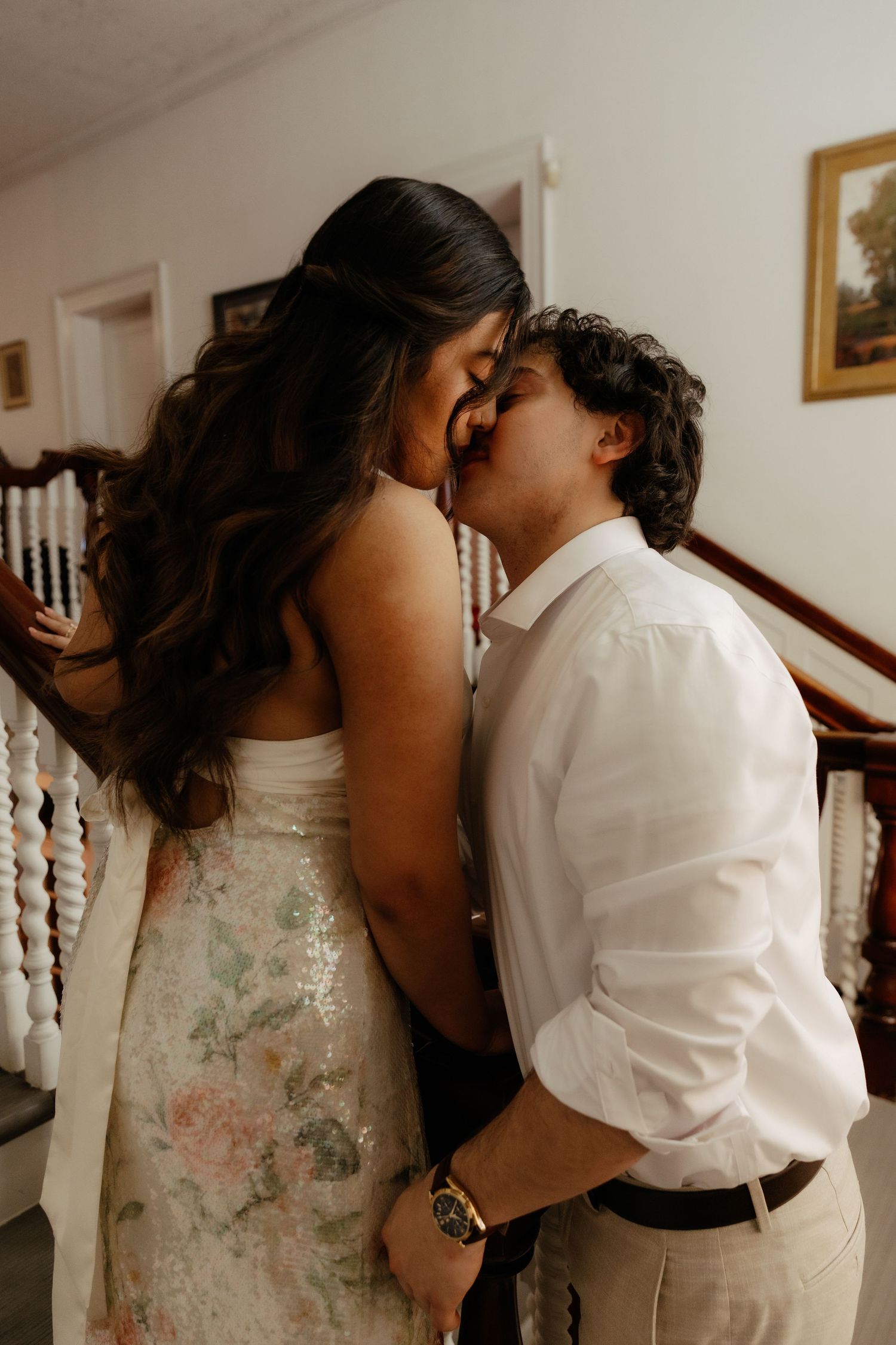 Couple shares a romantic kiss near staircase, bride in floral dress embracing groom in white shirt.
