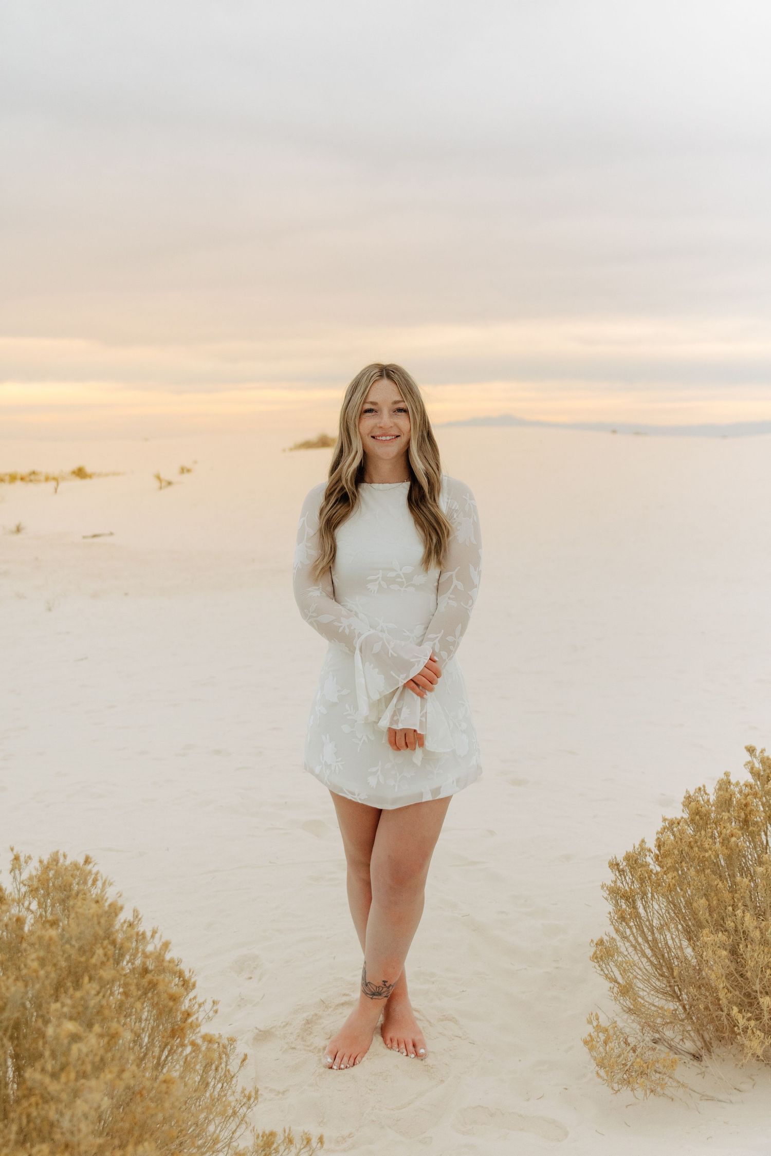 Barefoot woman in white floral lace dress standing on White Sands dunes at sunset.
