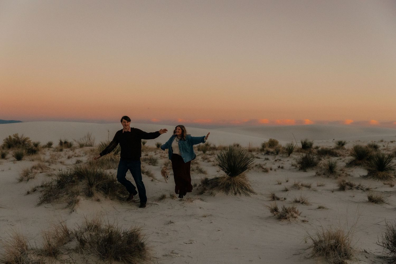 Joyful couple dancing and holding hands on white sand dunes surrounded by desert plants at dusk.