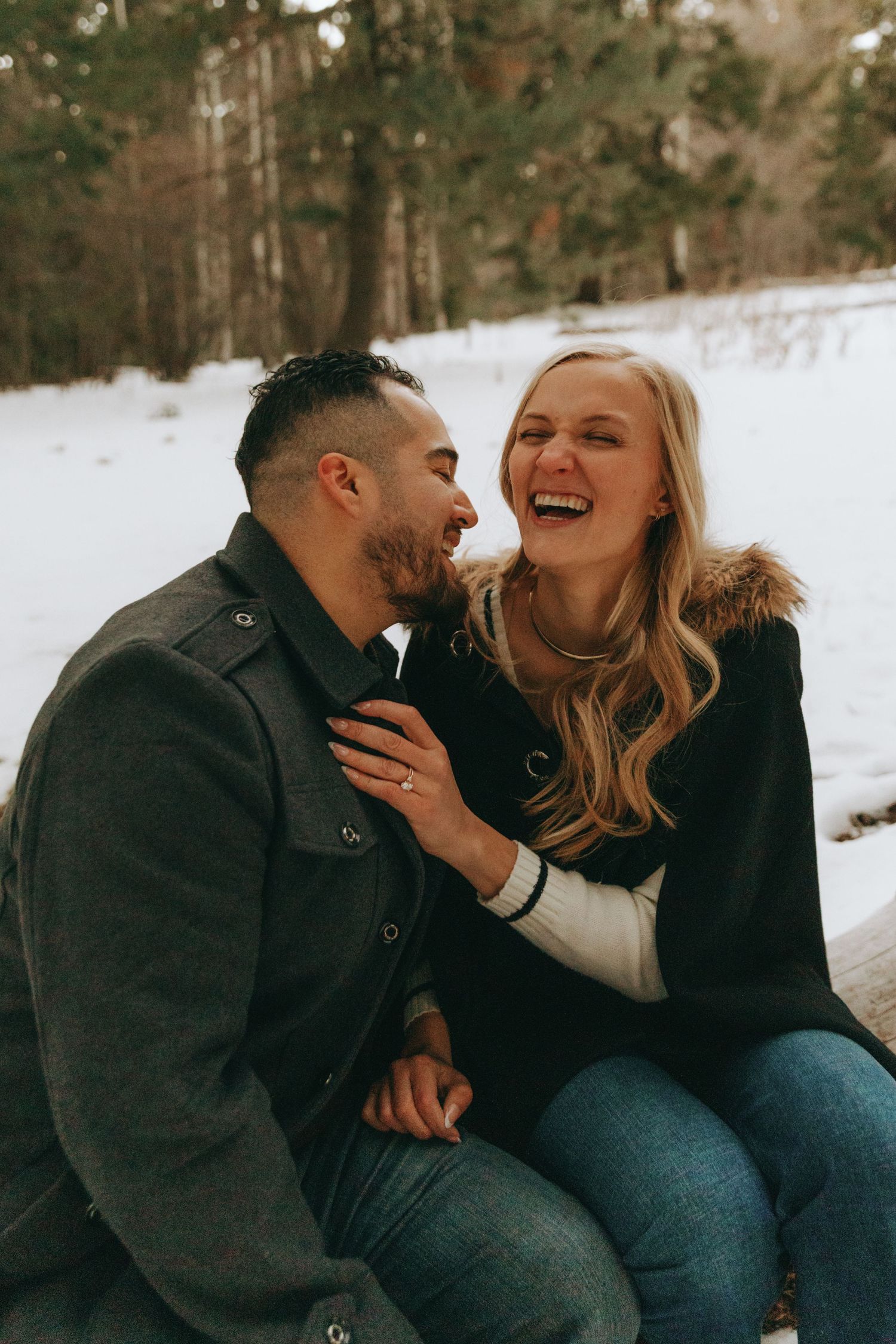 A laughing couple in winter coats shares a joyful moment outdoors in a snowy forest setting.