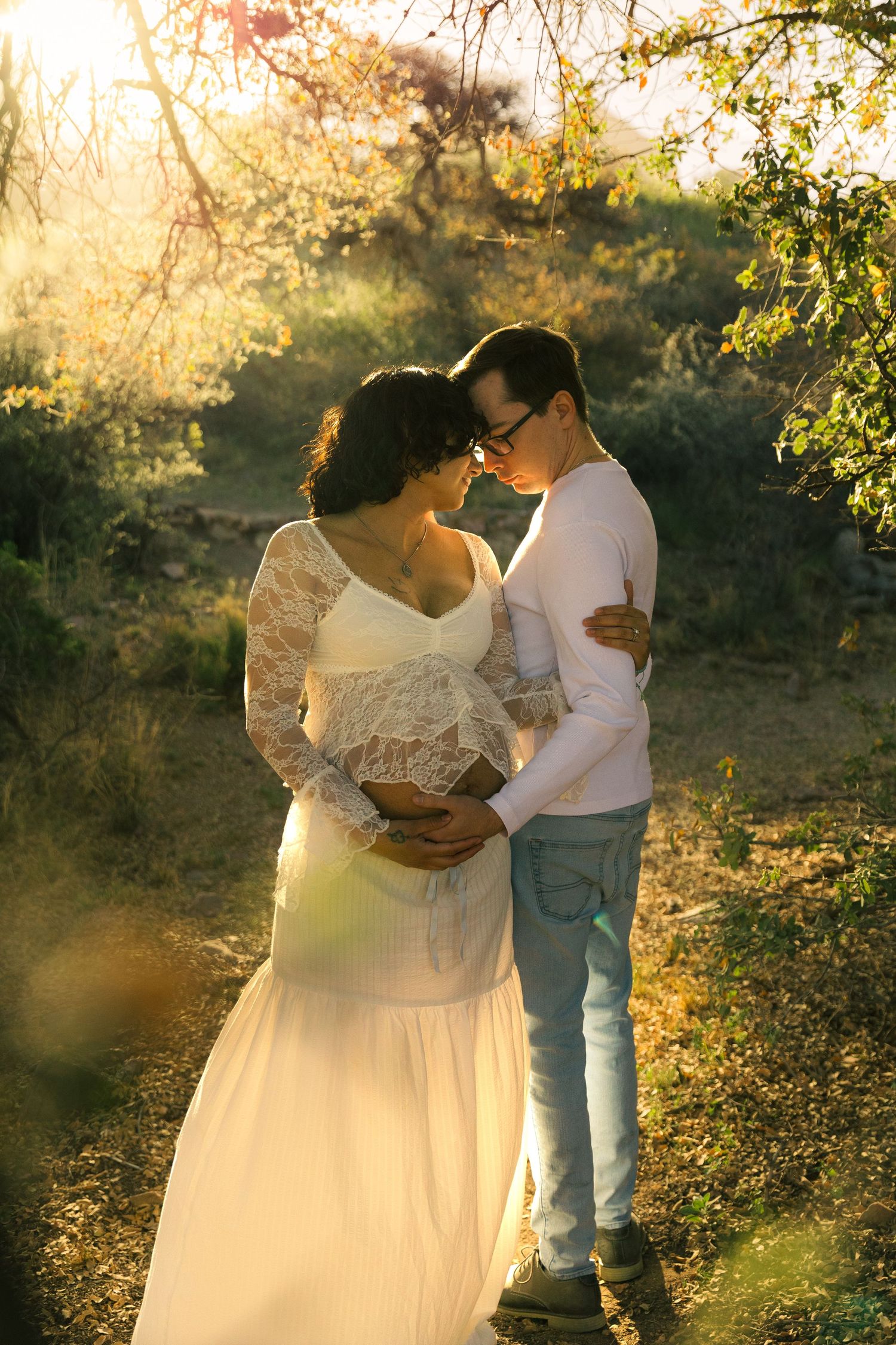 Couple embracing tenderly in golden sunset light among autumn trees, woman in white lace dress, maternity photo session.