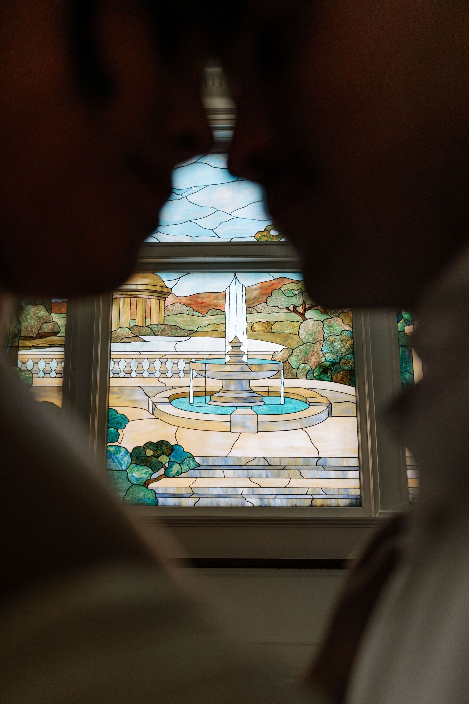 Stained glass window depicting a classical fountain garden scene with columns, trees, and blue sky.