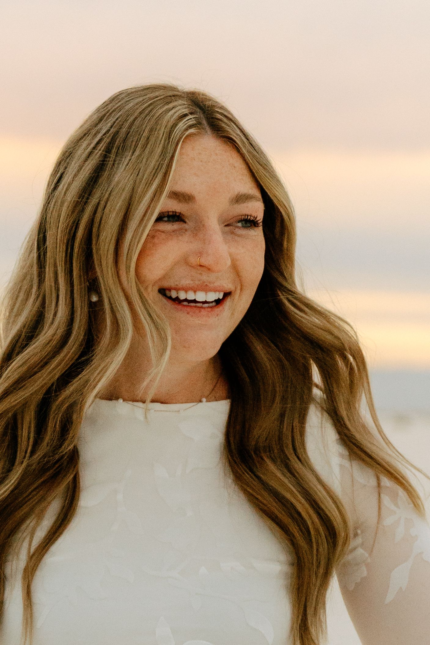 Smiling young woman with long blonde wavy hair wearing a white lace dress at sunset on a sandy beach.