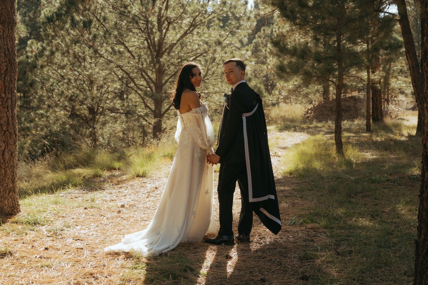Bride in white gown and groom in black cloak hold hands on a sunlit forest path among pine trees.