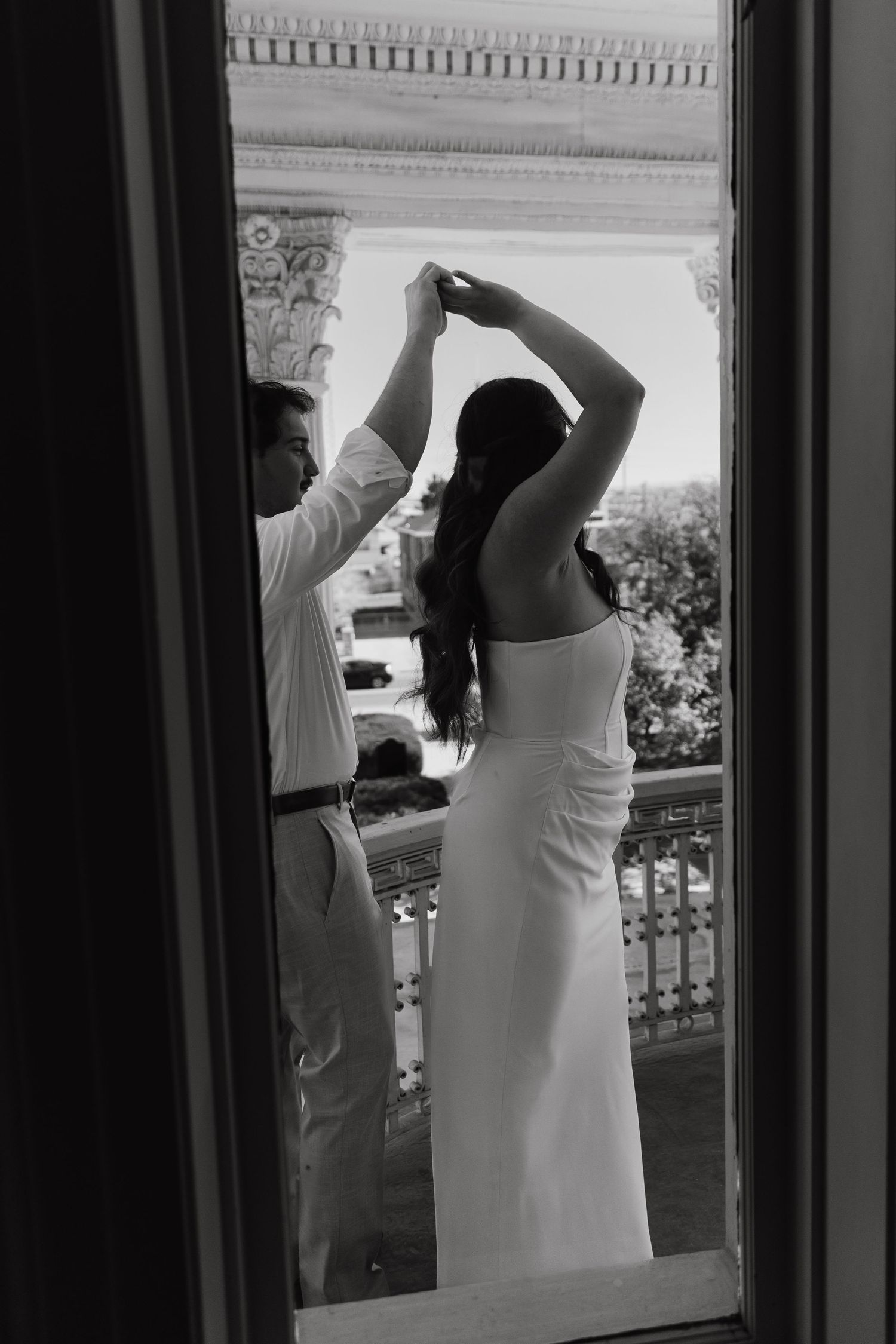 Black and white photo of a couple on a balcony, groom helping bride with her hair, wearing elegant wedding attire.