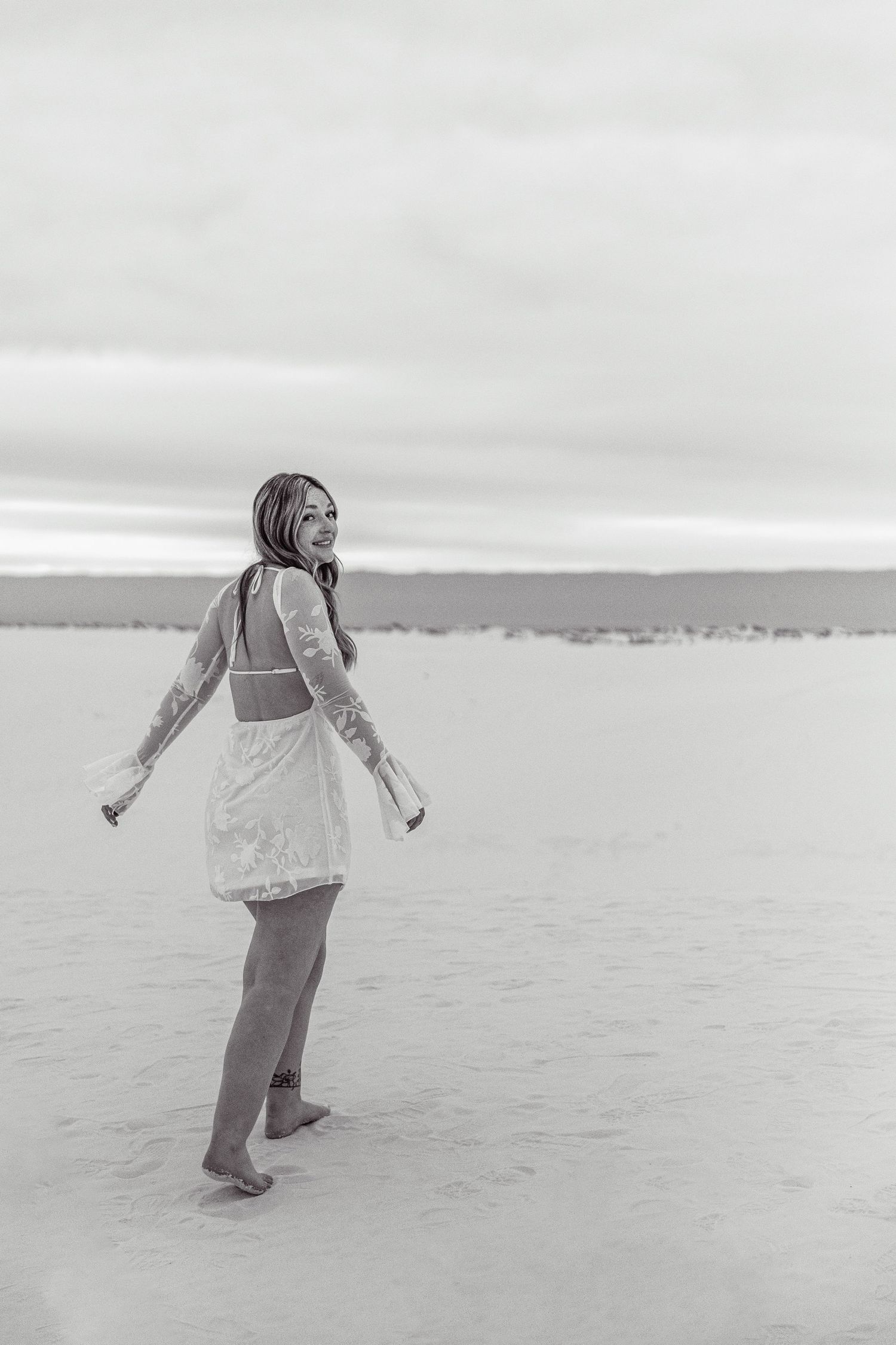 Black and white photo of a young woman in a white lace dress looking back over her shoulder on a sandy beach.