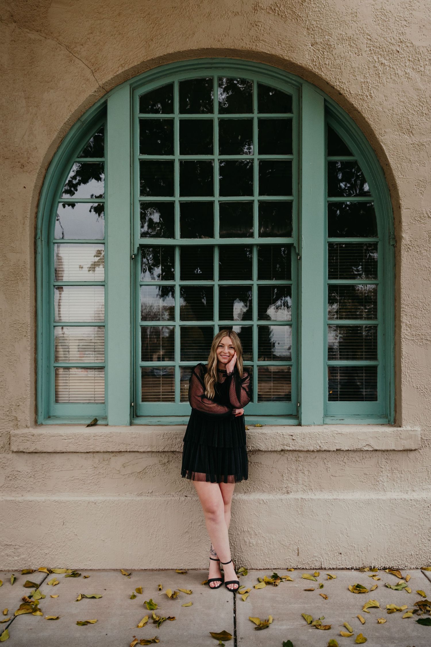 Blonde woman in black dress posing in front of large arched teal window.