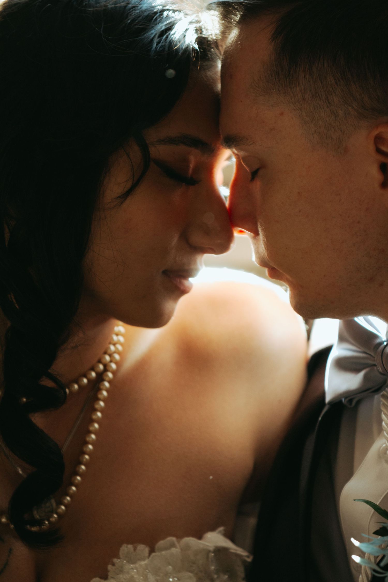 A bride and groom share an intimate moment, foreheads touching, as soft light illuminates their wedding attire and pearl necklace.