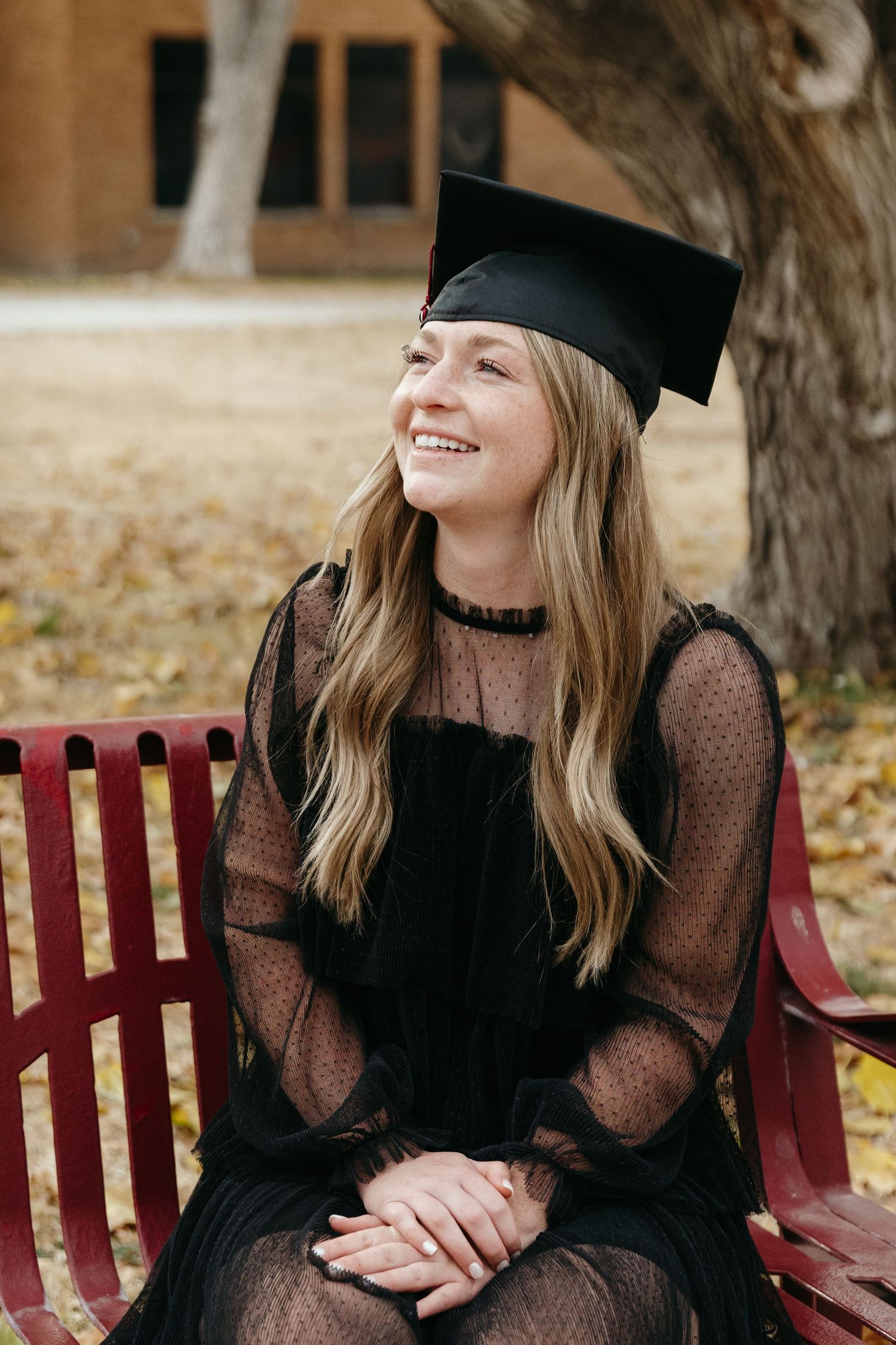 Smiling graduate in black lace dress and mortarboard cap sitting on red bench outdoors.