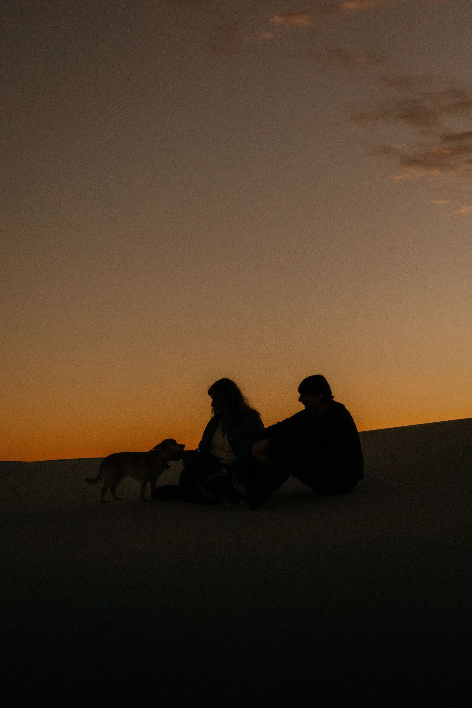 Silhouette of couple and dog sitting on sand dunes at sunset.