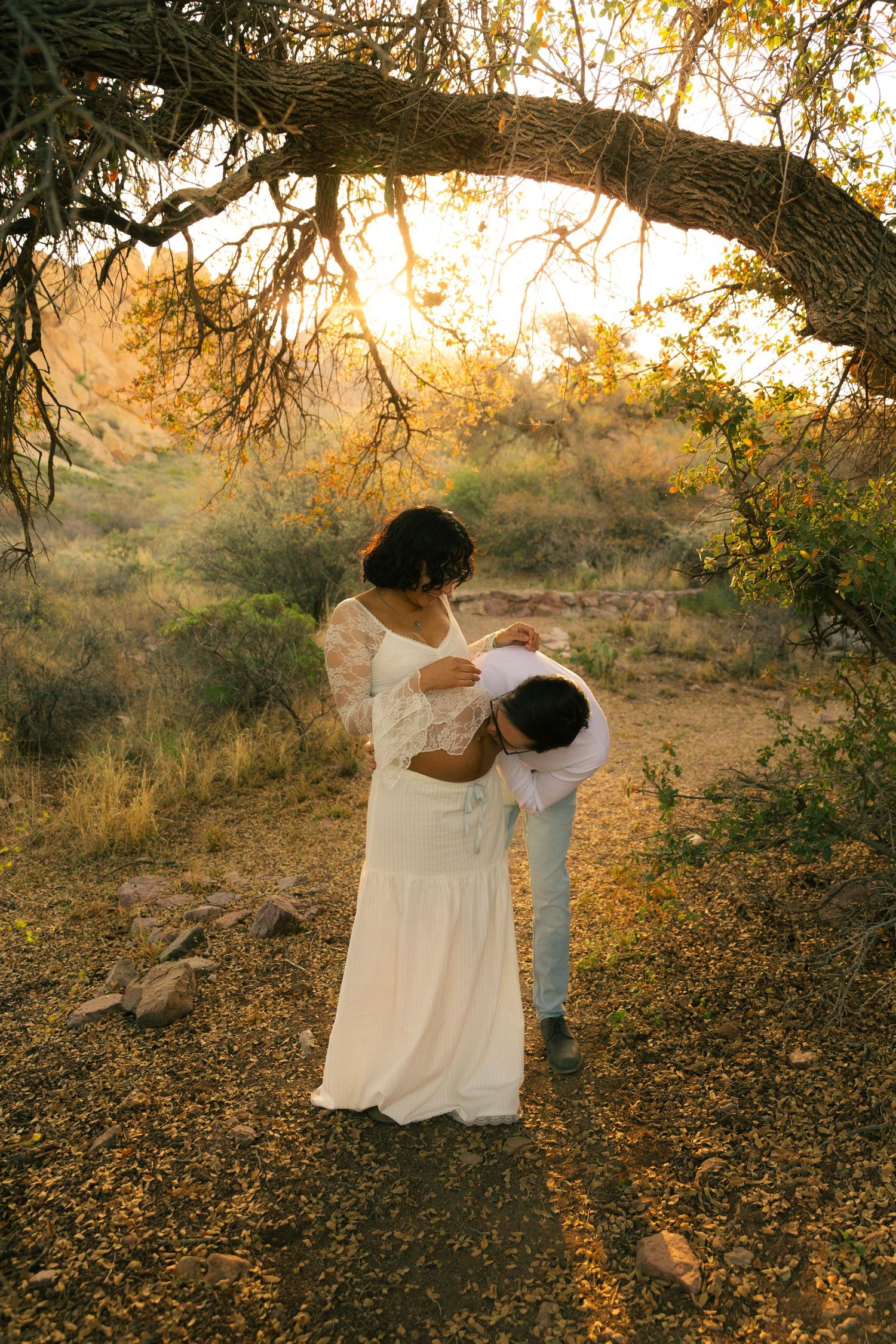 Couple sharing tender moment under arching tree branch in golden sunset light, woman in white lace maternity gown outdoors.