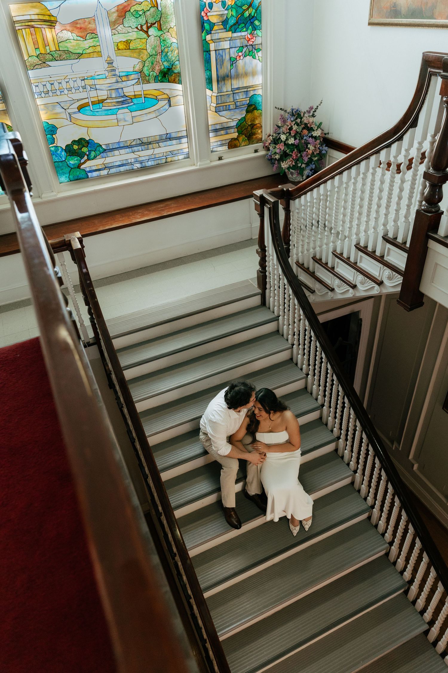 Bride and groom kissing on grand staircase with stained glass window in elegant historic venue.