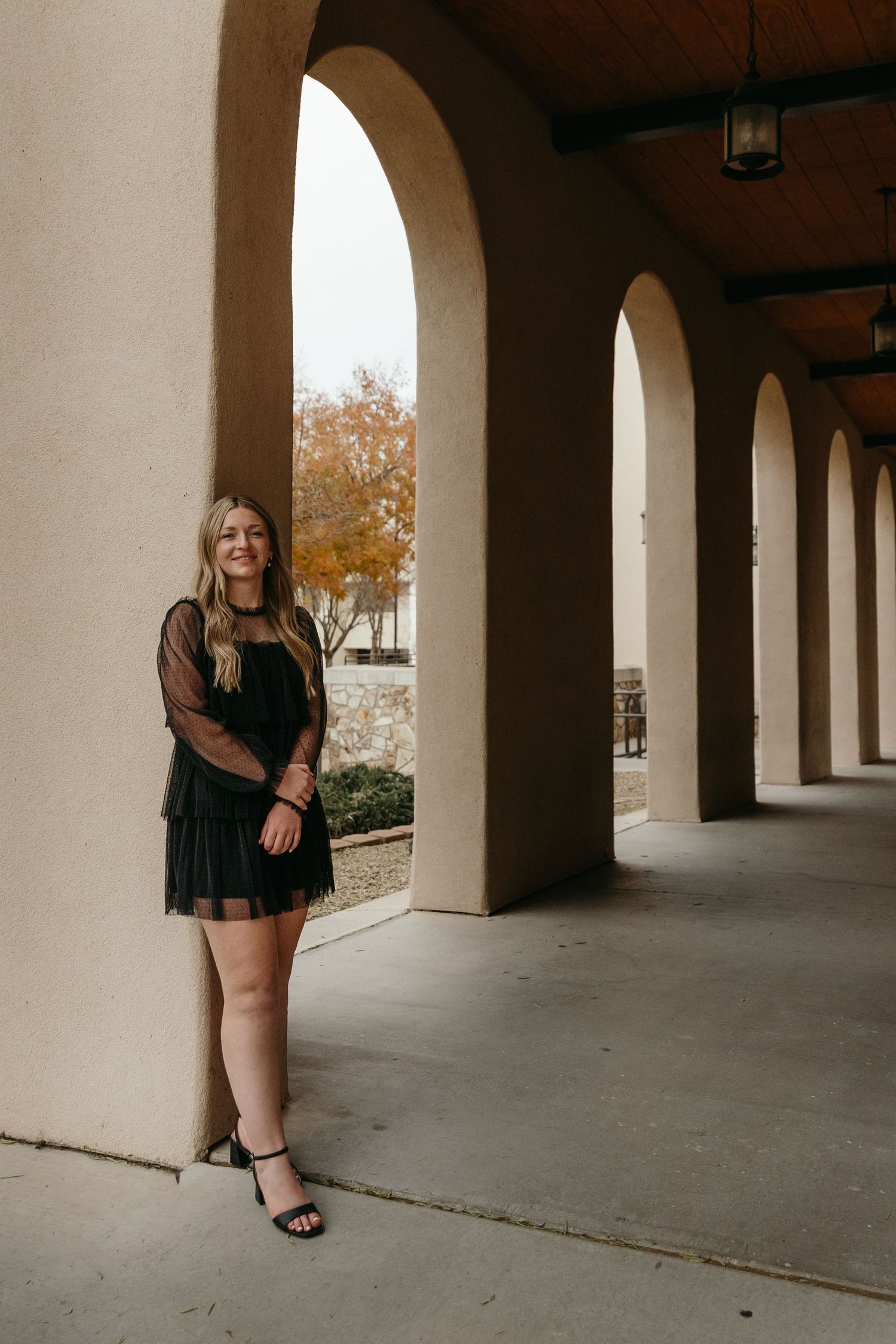 Young woman in black dress posing under arched colonnade of university building.