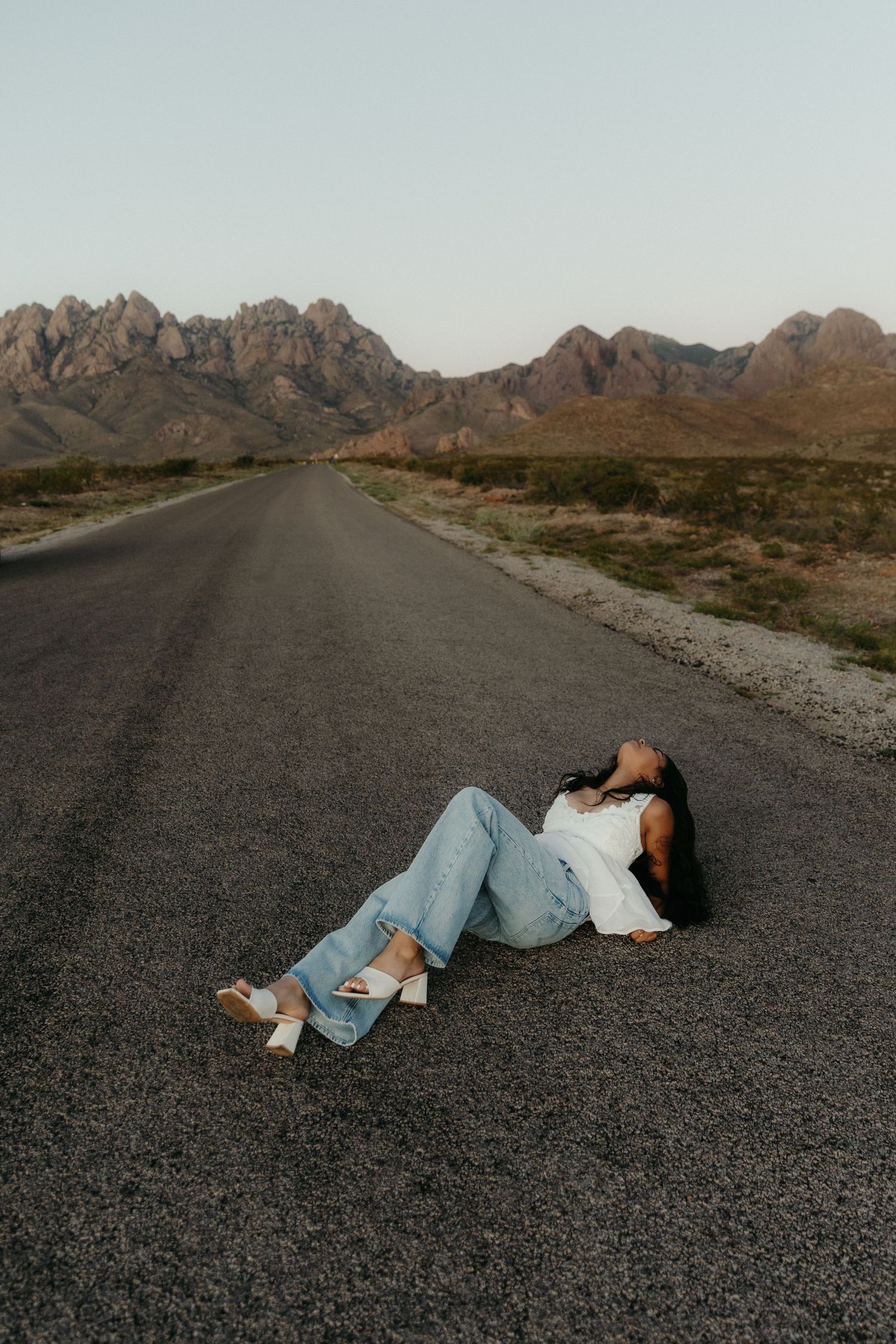 Woman in white top and jeans lying on a desert road with dramatic mountain backdrop at dusk.