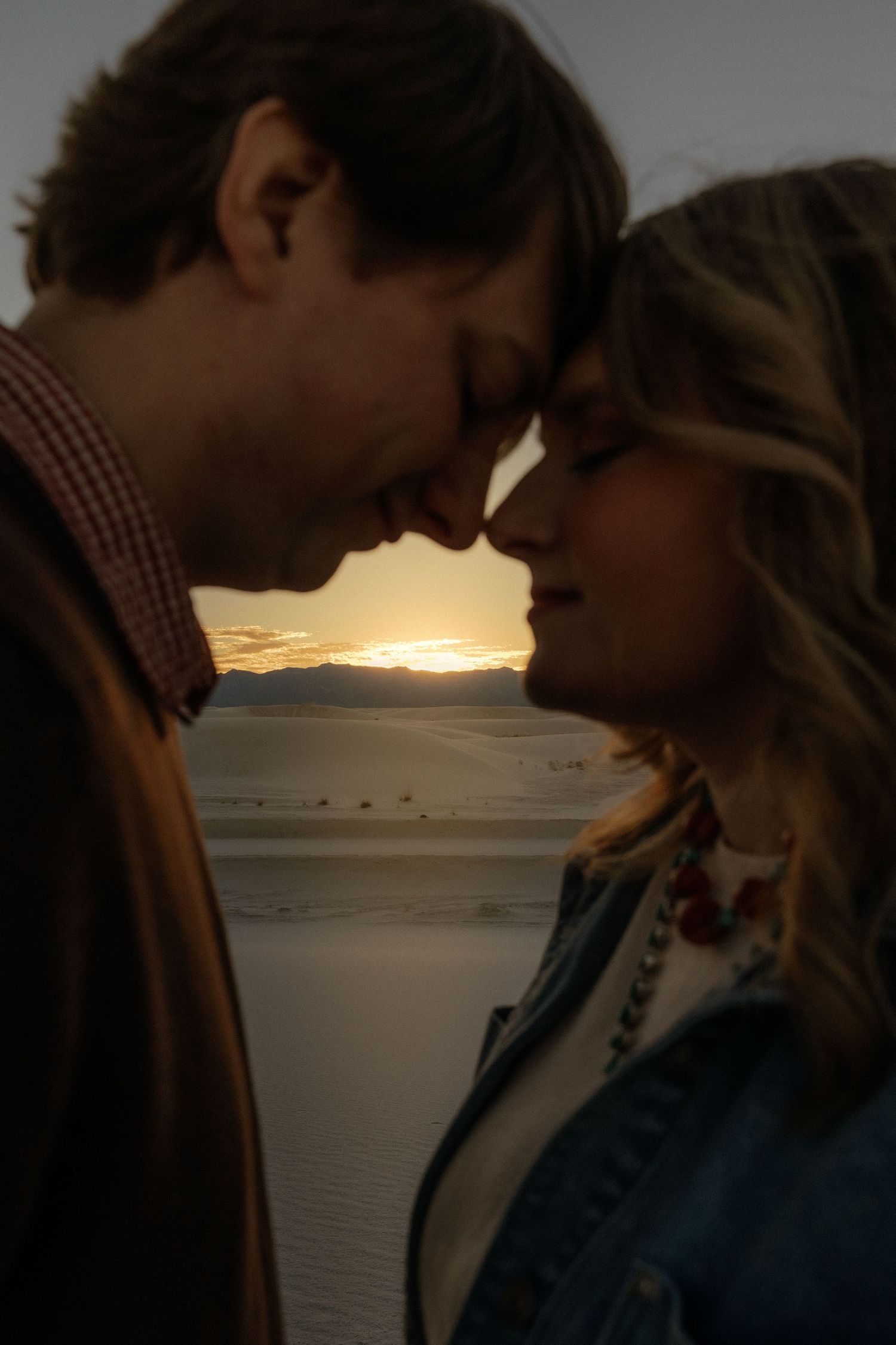 Romantic couple touching foreheads at sunset over white sand dunes.