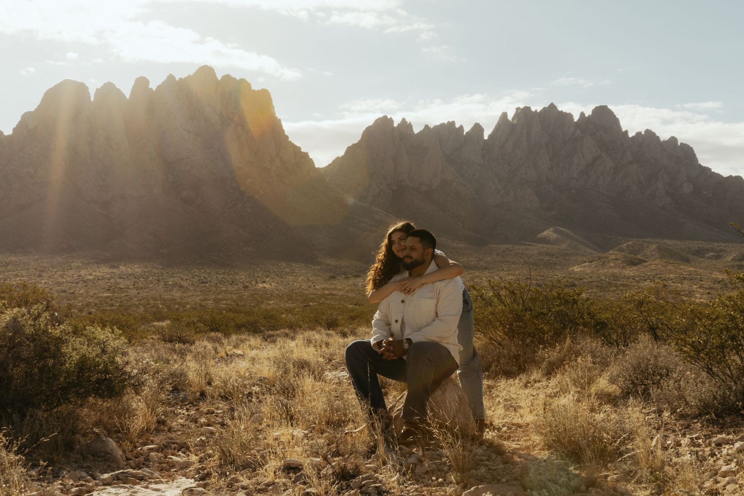 Couple embracing in golden desert landscape with dramatic rocky mountains and sunburst in background.