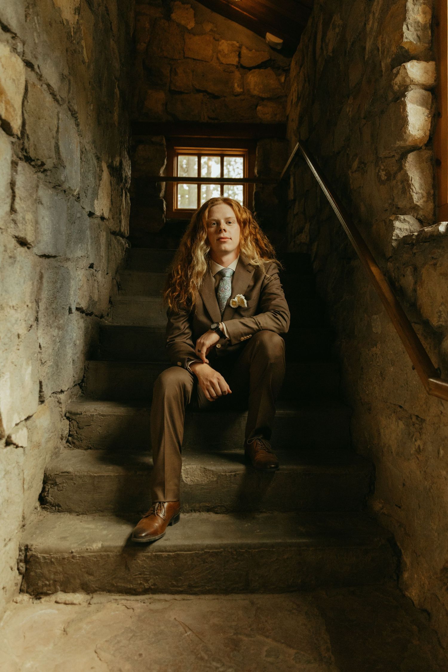 Redhead person in brown suit sitting on stone staircase with window light, wearing boutonniere and tie.