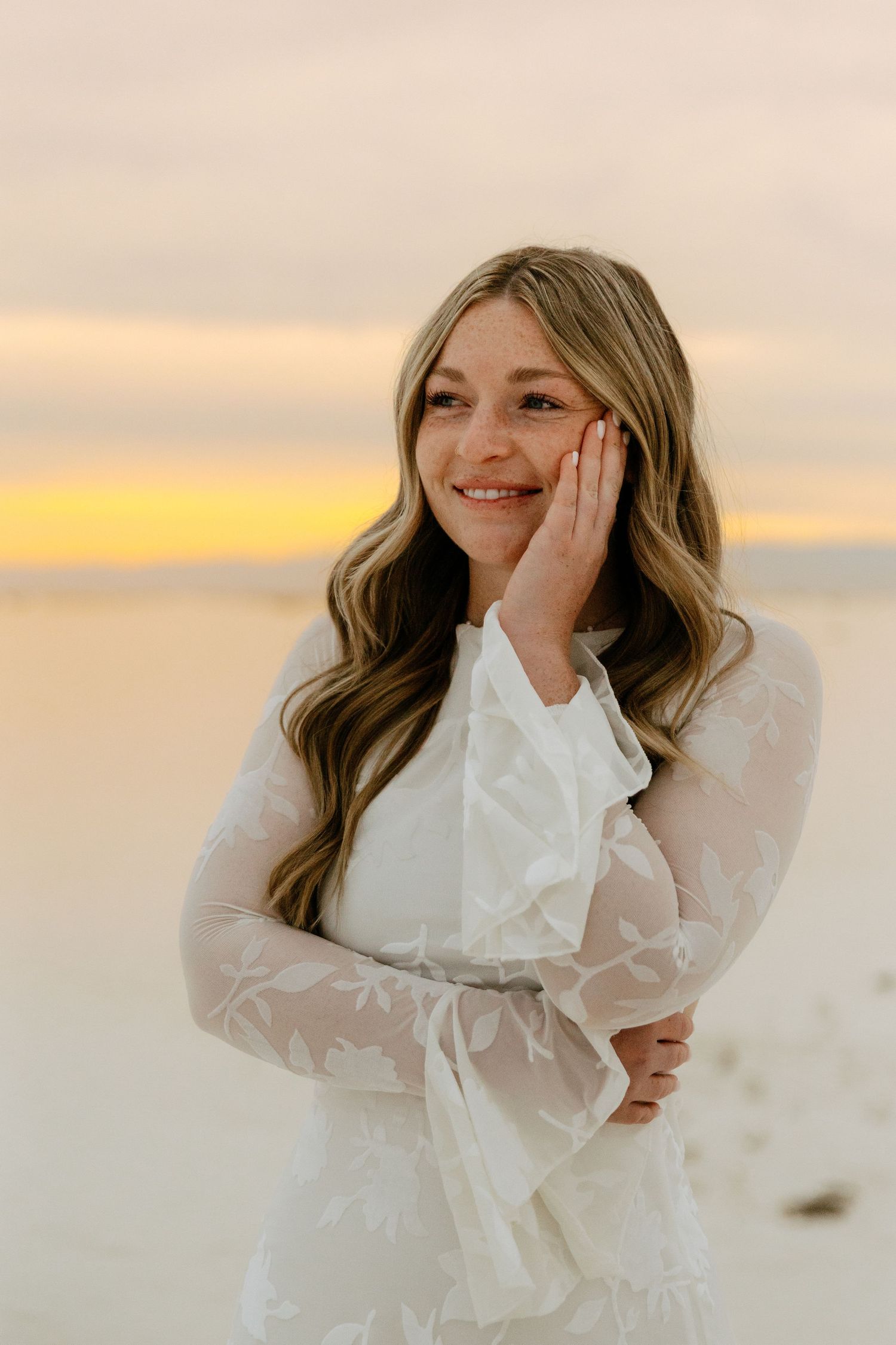 Smiling blonde woman with freckles in white lace dress rests hand on cheek at beach sunset.