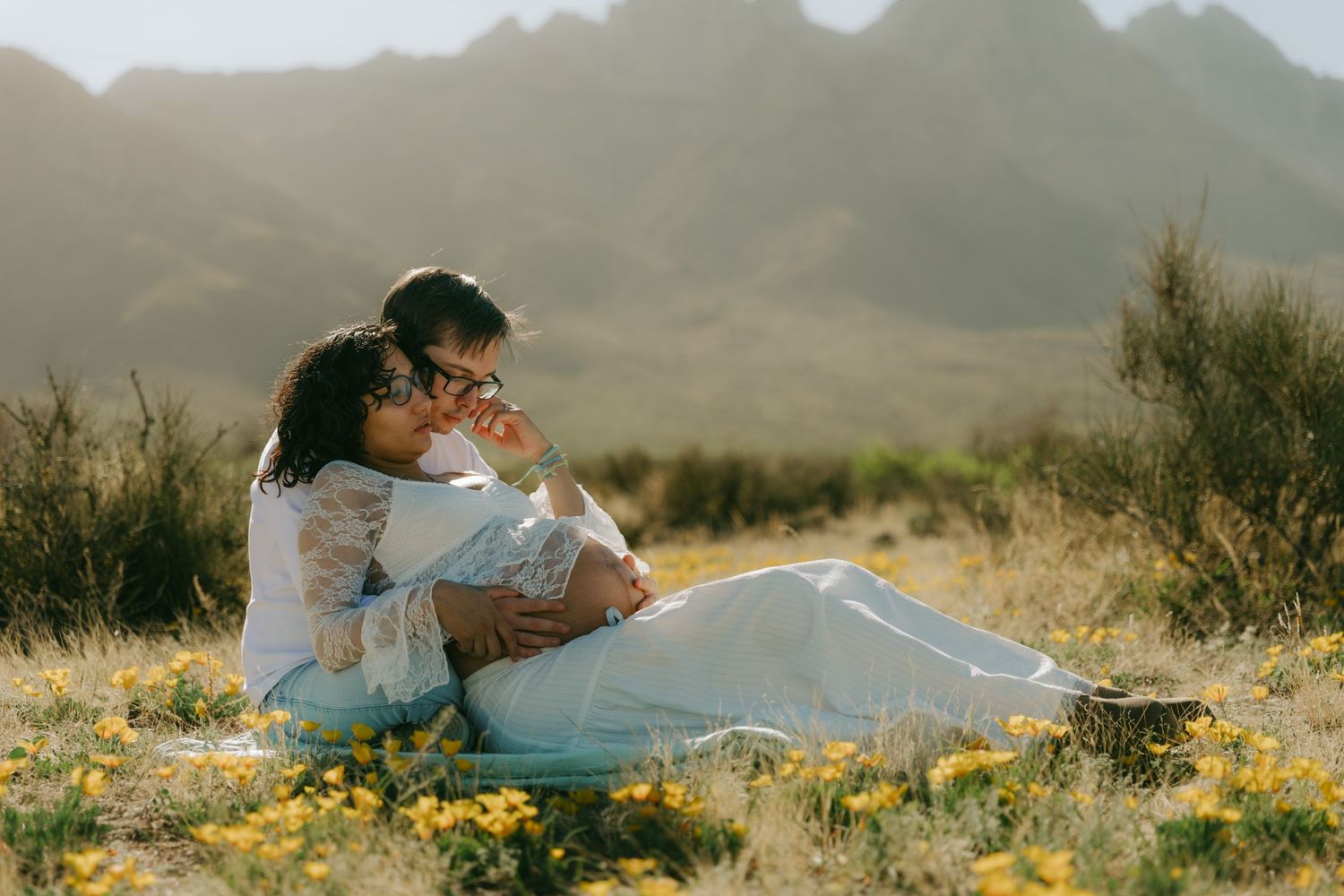 Couple sitting together in a sunny field of yellow wildflowers with mountains in the background.