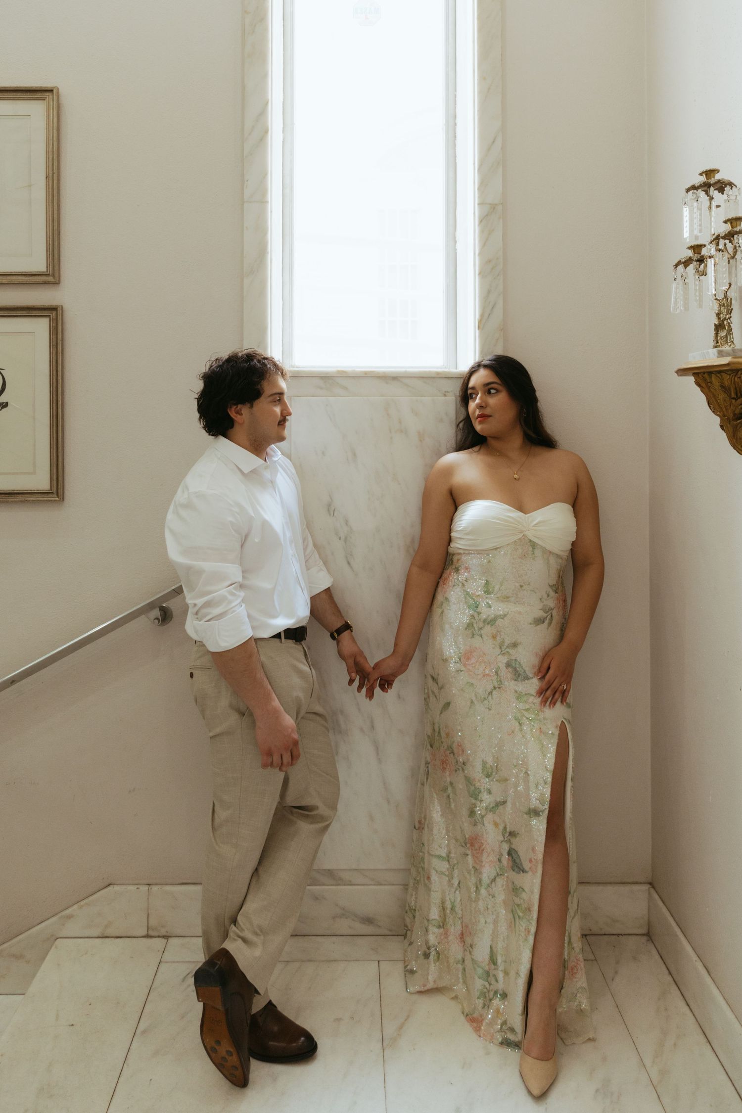 Couple holding hands by marble wall and window, woman in floral gown, man in white shirt and tan pants.