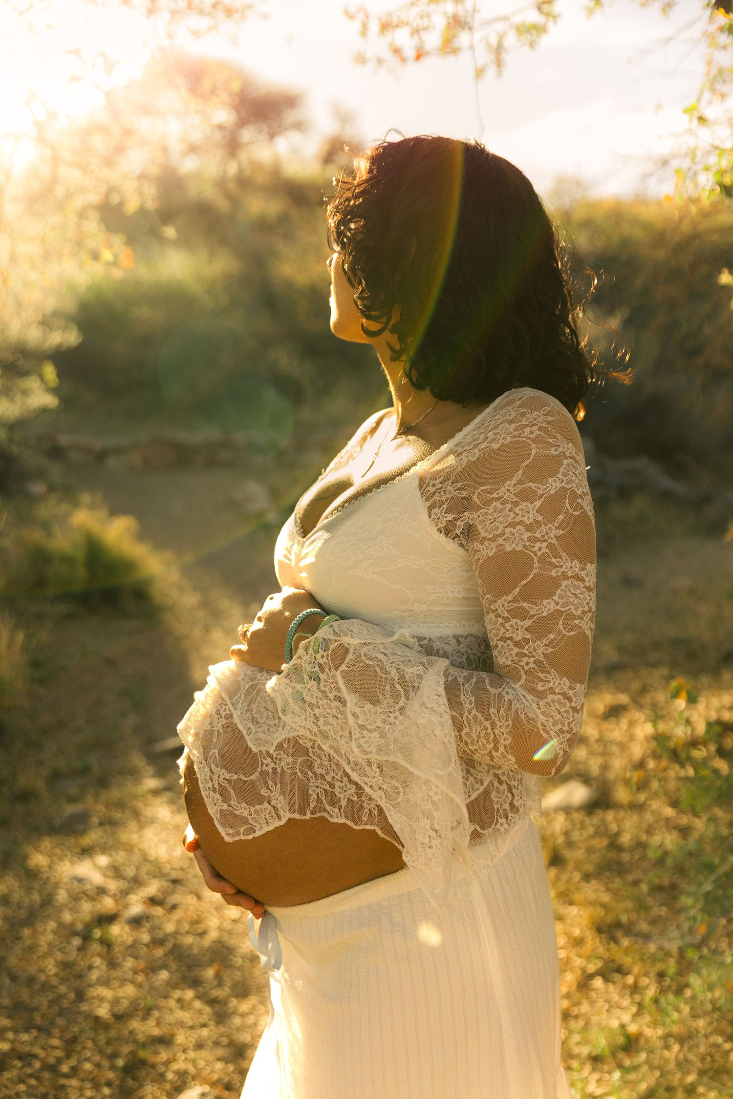 Pregnant woman in white lace dress cradling baby bump outdoors in golden sunlight during maternity photo shoot.