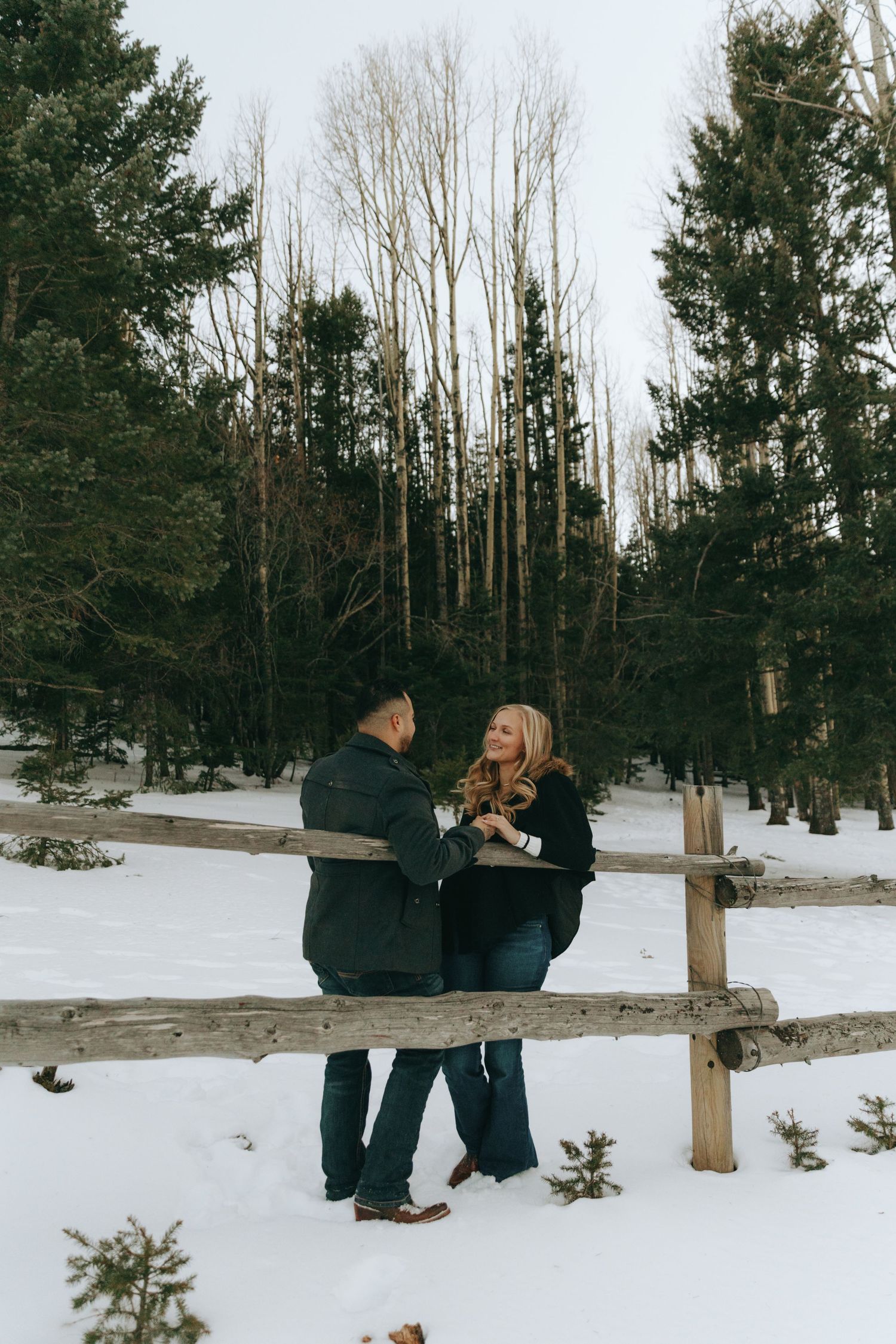Couple holding hands and smiling in a snowy winter forest with wooden fence and evergreen trees.