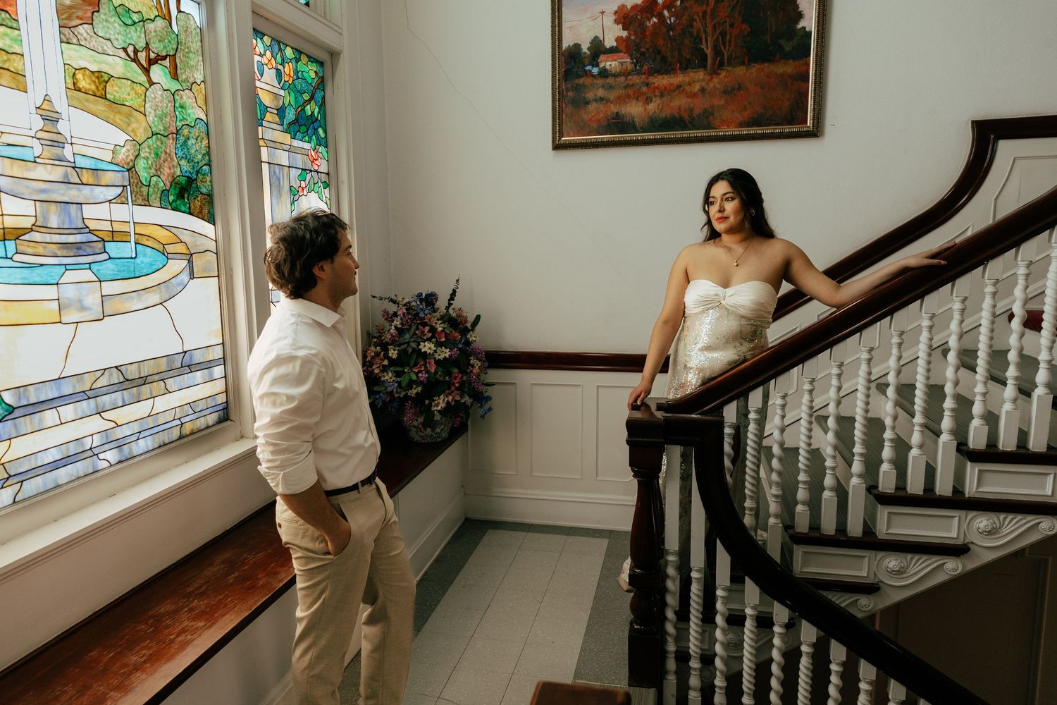 Bride in strapless gown posing on staircase while groom admires her near stained glass window.