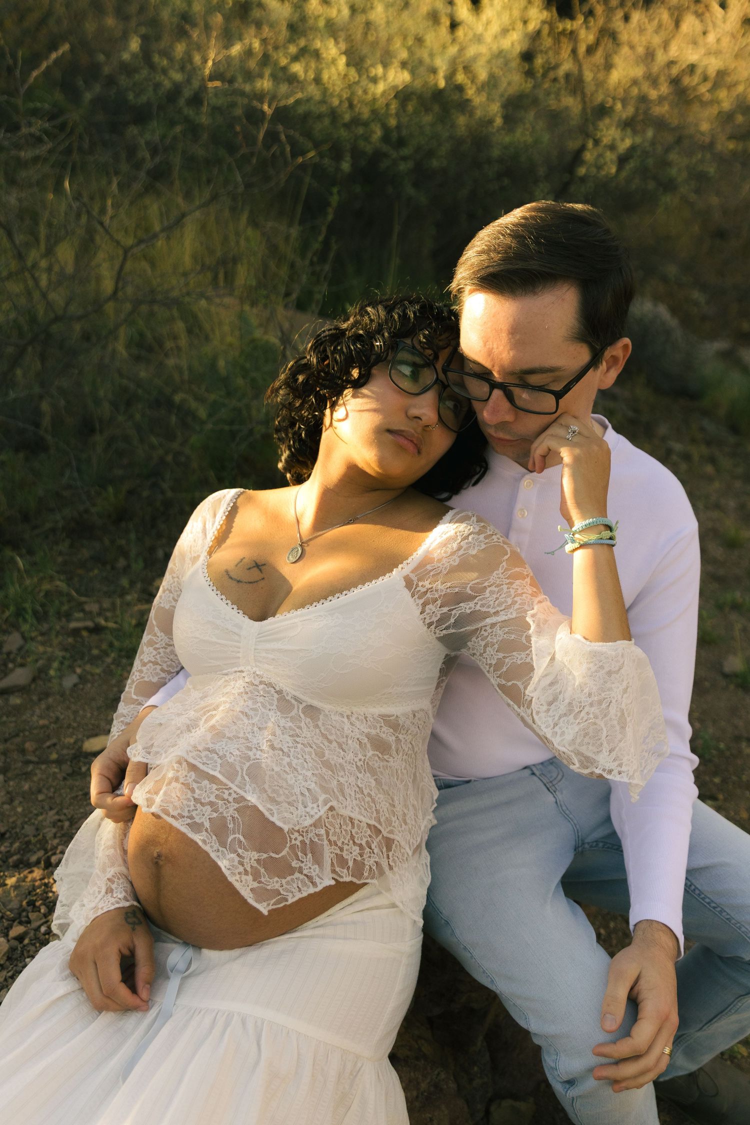 Pregnant woman in white lace dress sits with partner in golden hour light, both sharing an intimate tender outdoor moment.